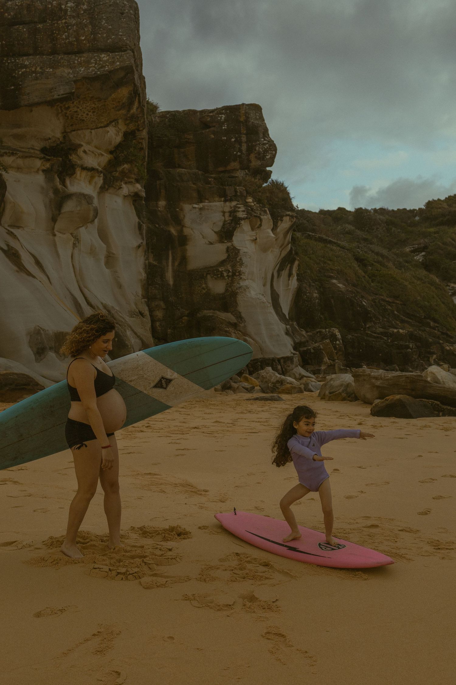 Pregnant woman holds surfboard while young girl practices surfing on pink board at sandy beach with rocky cliffs.