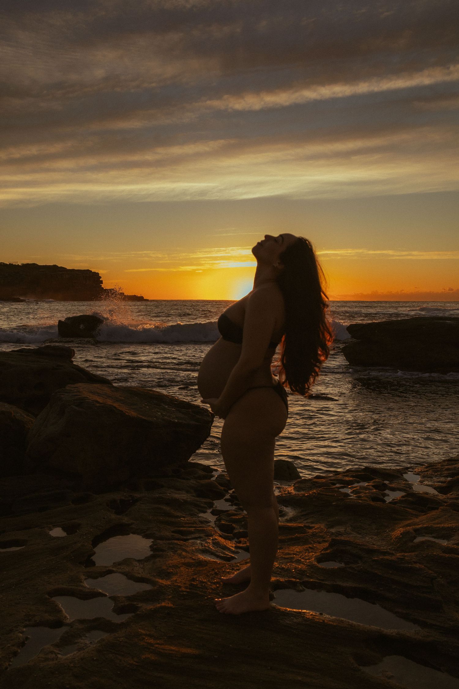 Pregnant woman silhouetted against a vibrant ocean sunset, standing on rocky shore in a bikini with hair flowing in the breeze.