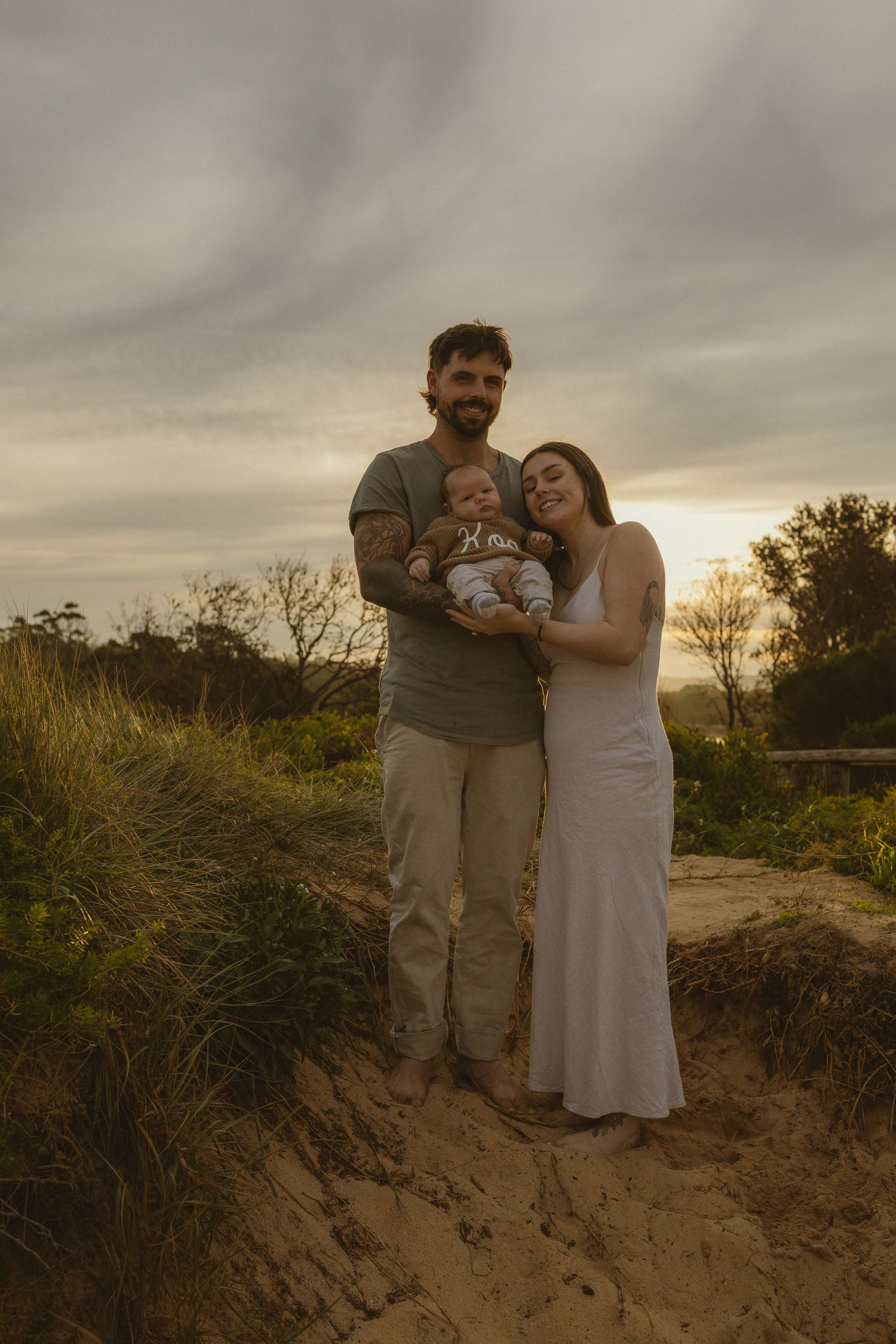 A couple smiles holding their newborn baby on a sandy path at golden hour with lush greenery and a cloudy sky.