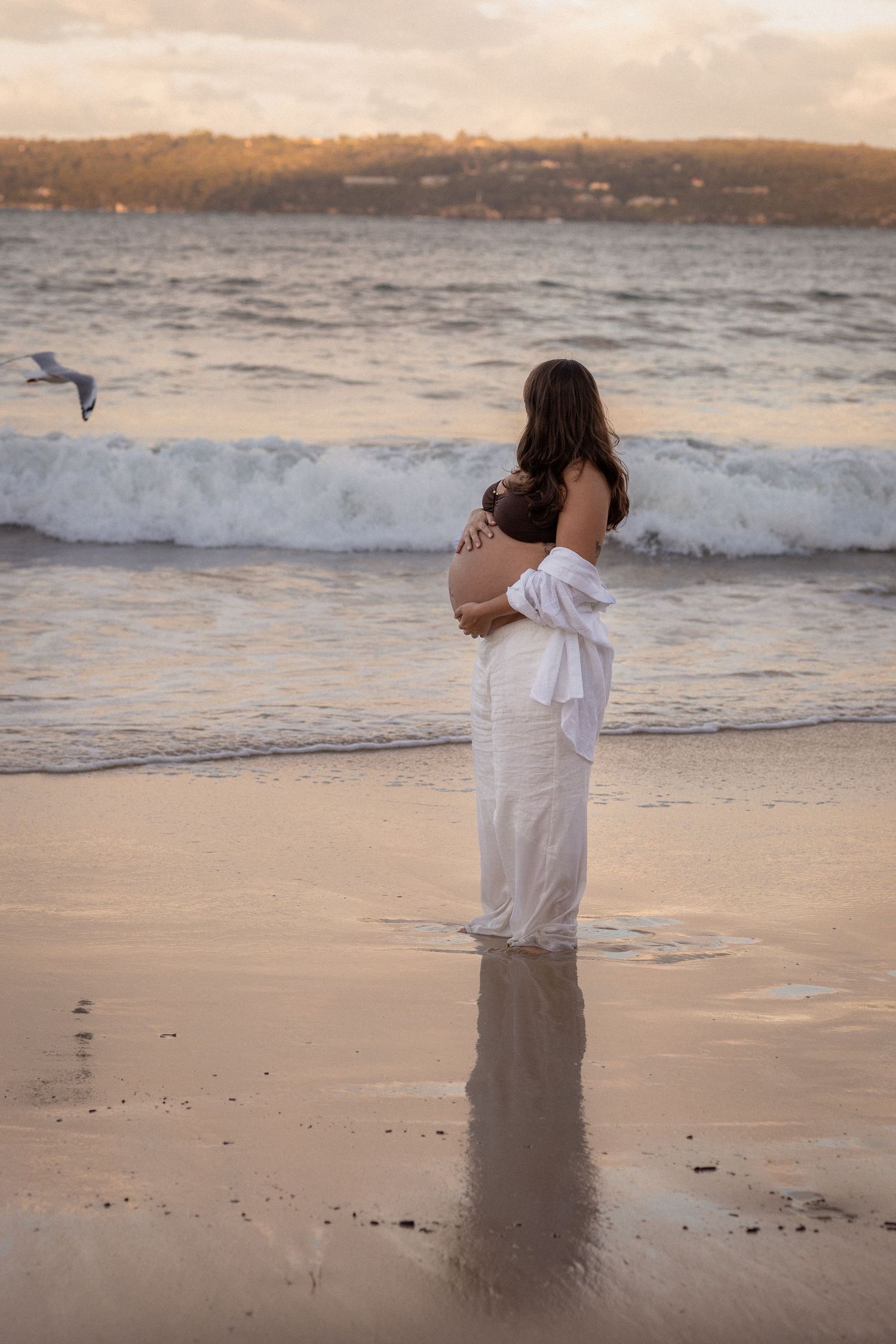 Person in white clothing stands in shallow ocean waves at sunset creating reflections in wet sand on beach.