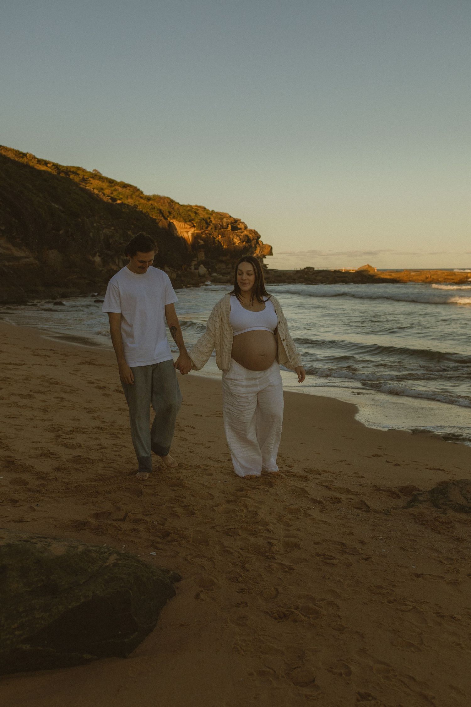 Pregnant couple walking hand in hand on a sandy beach at sunset, with rocky cliffs and ocean waves in the background.