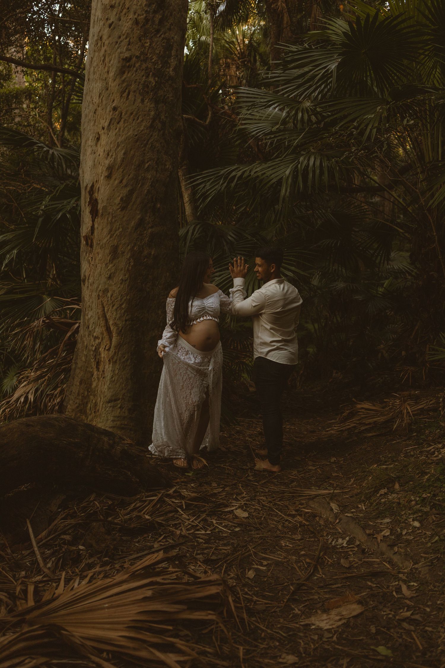 Pregnant woman in white dress leaning against large tree trunk with partner beside her in tropical forest.