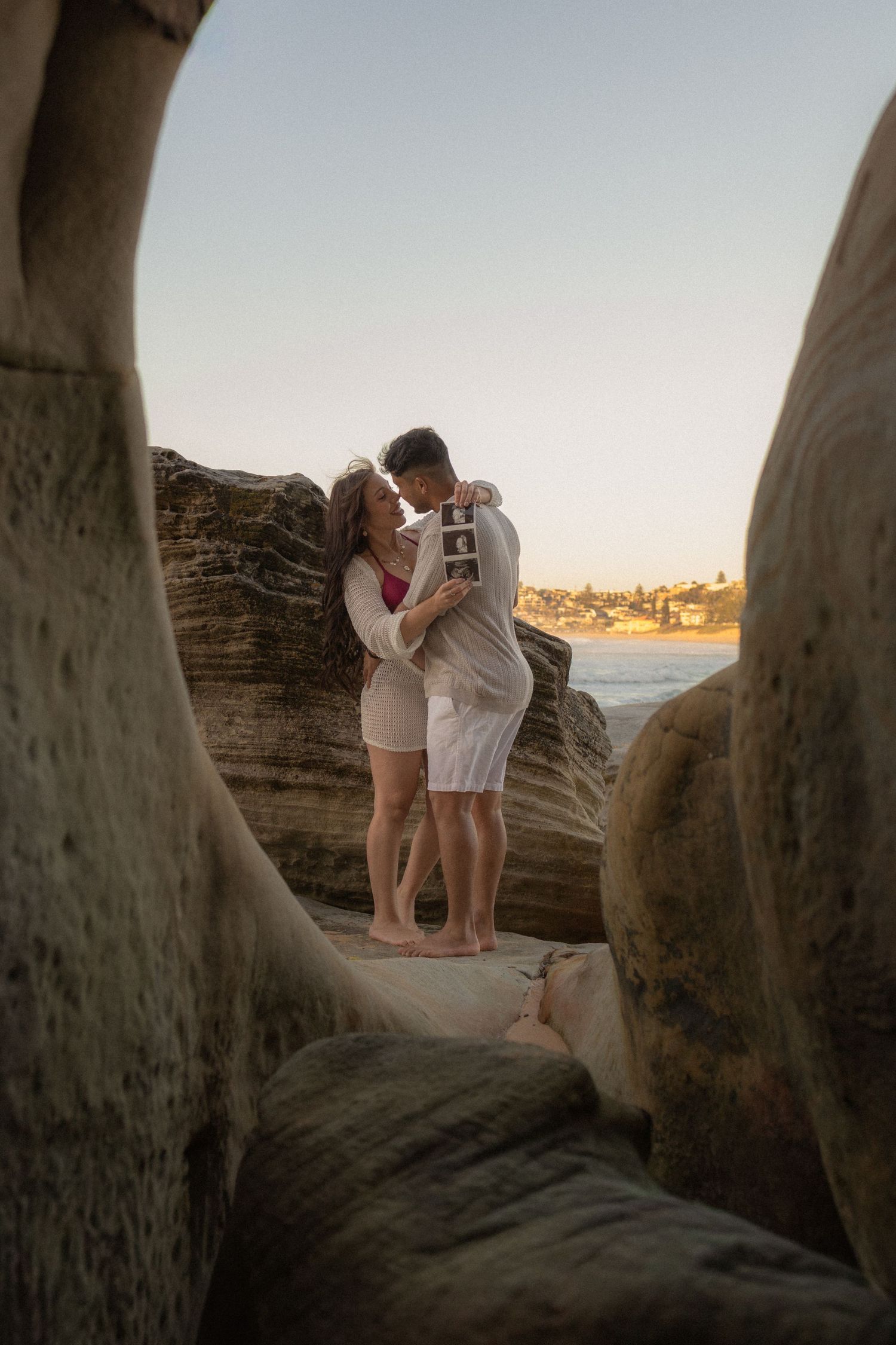 Couple embracing and kissing on rocky coastal cliffs at sunset, holding ultrasound photos to announce pregnancy.