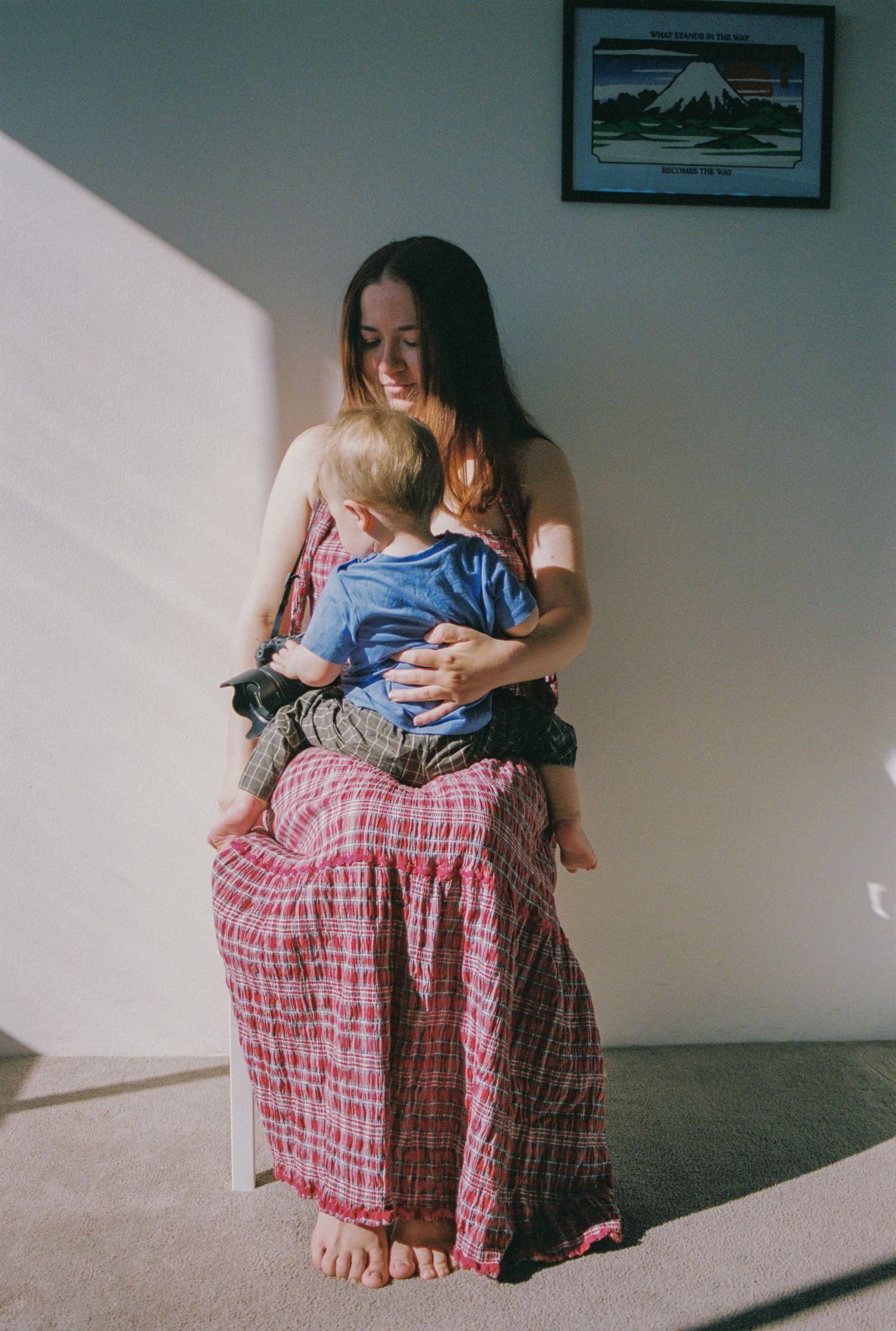 A woman in a red plaid dress sits barefoot holding a toddler, smiling warmly in sunlit room with framed art.