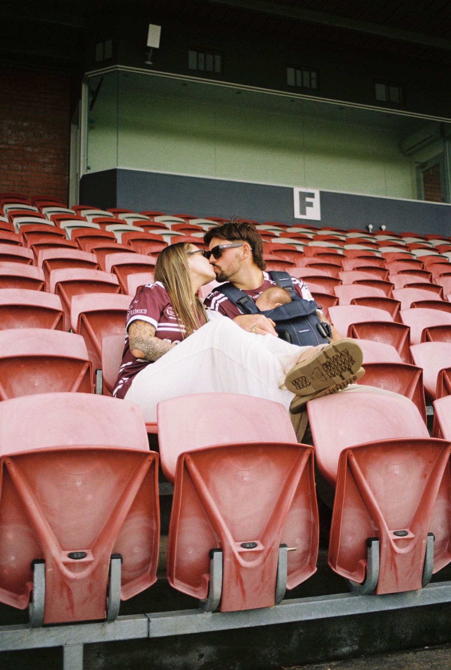 Couple sitting close together in red stadium seats, wearing sports jerseys, sharing a romantic moment in an empty grandstand.