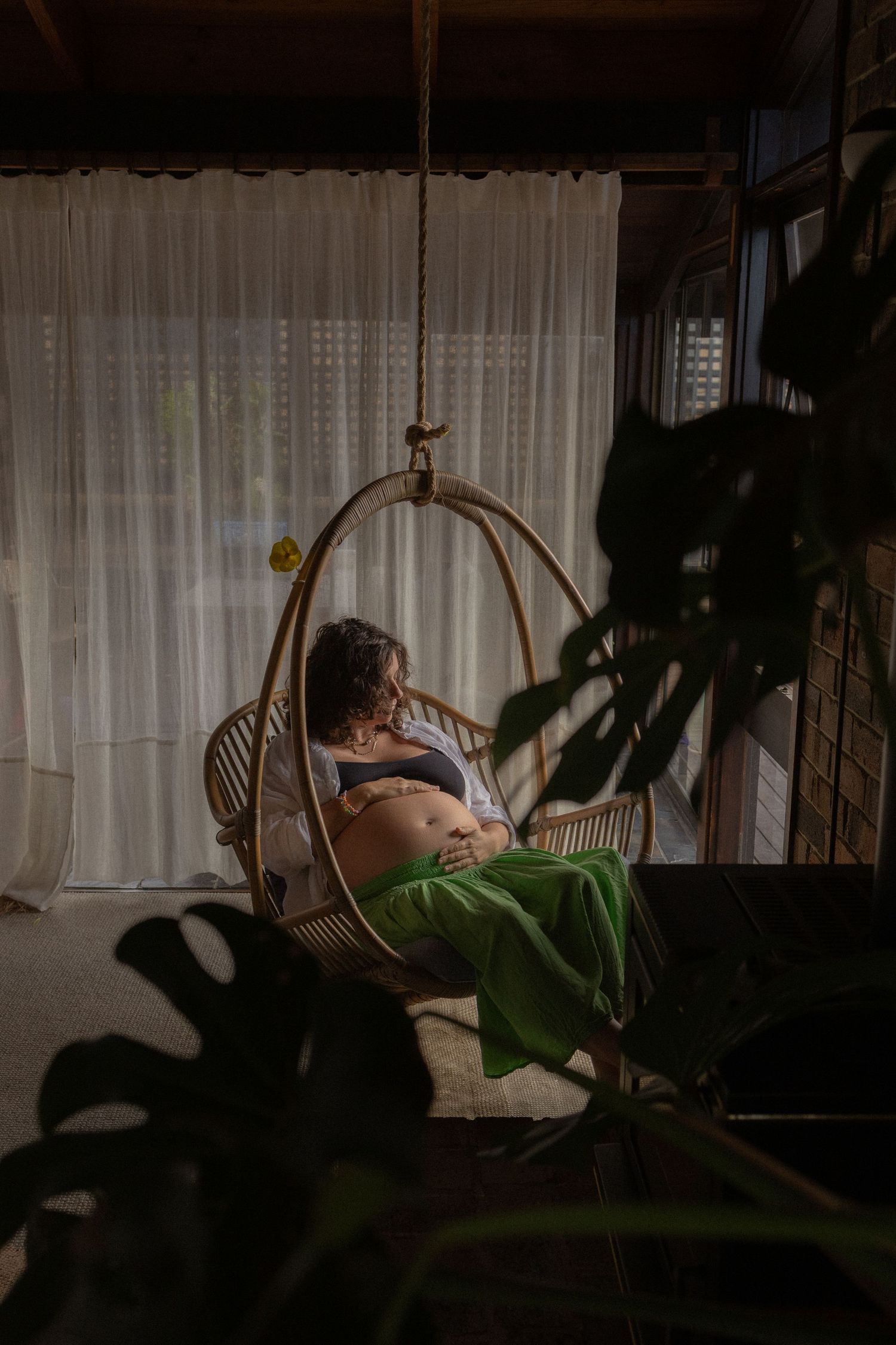 Pregnant woman relaxing in a rattan hanging chair by sheer curtains, cradling her baby bump in warm natural light.
