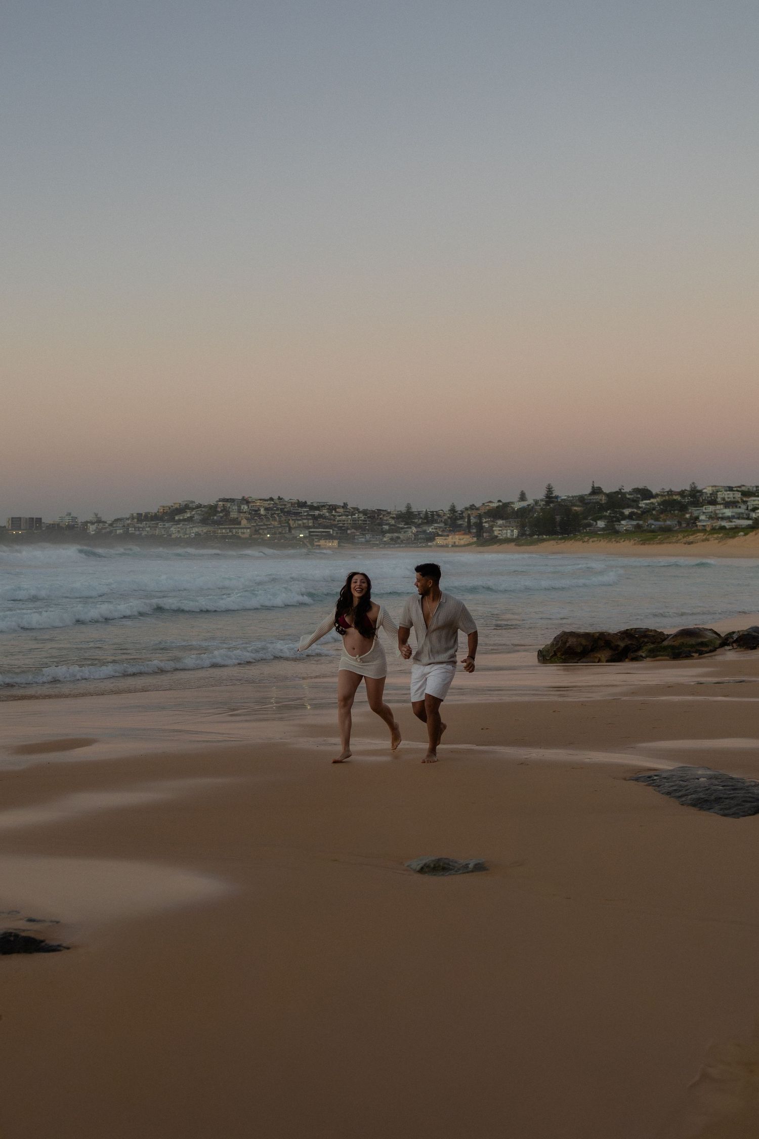 A couple runs hand-in-hand along a sandy beach at sunset with ocean waves and a coastal town in the background.