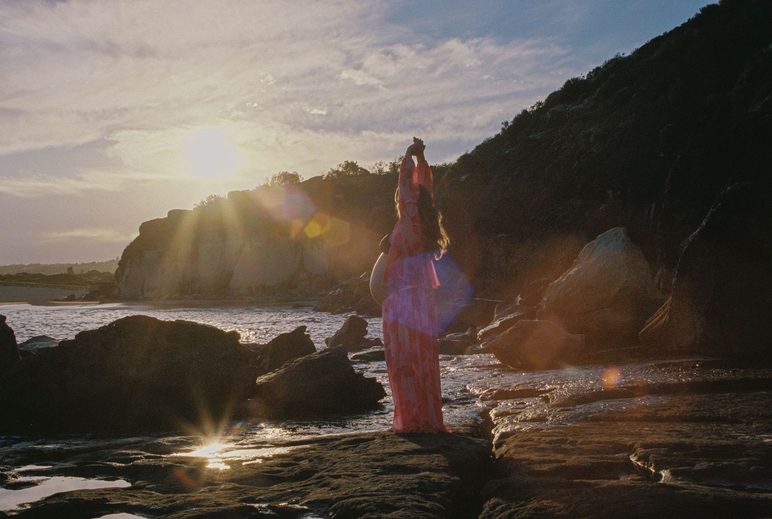 Woman in pink dress standing on rocky beach at sunset with golden light and ocean waves.
