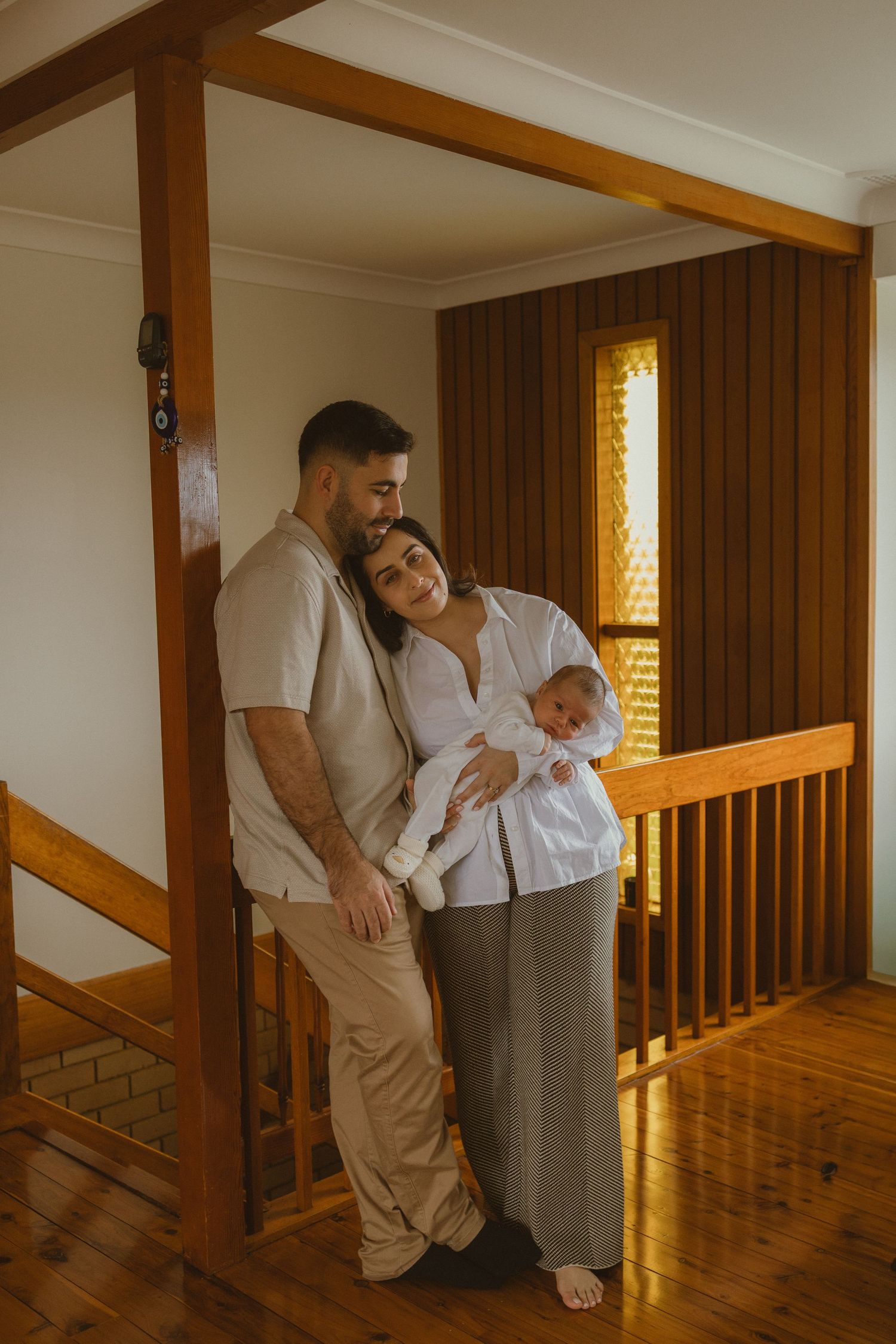 A couple stands together holding their newborn baby in a warm wooden home interior.