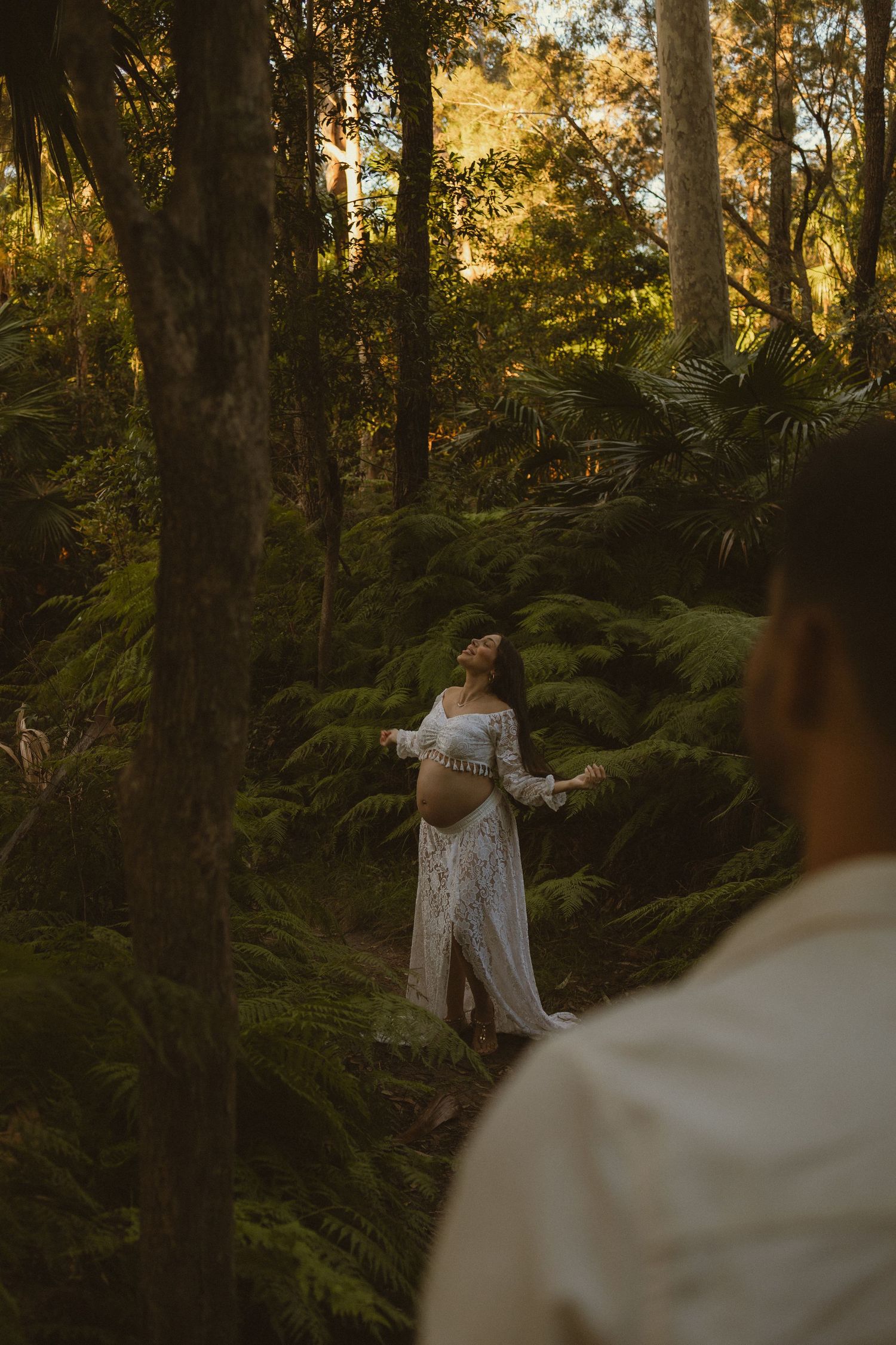 Pregnant woman in white lace dress standing among lush forest ferns in golden sunlight.
