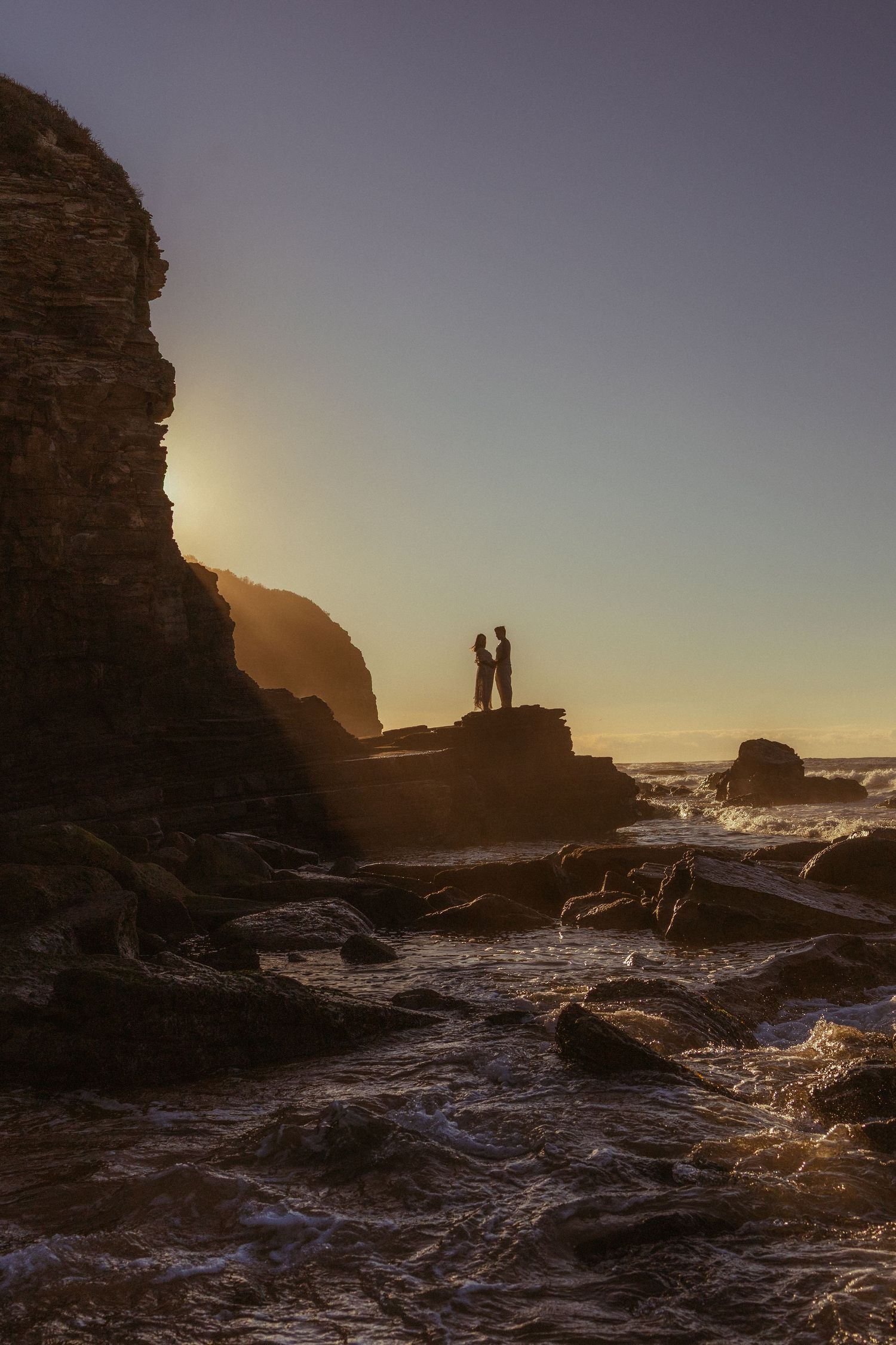Silhouette of couple standing on rocky coastal cliffs at golden sunset with ocean waves below.