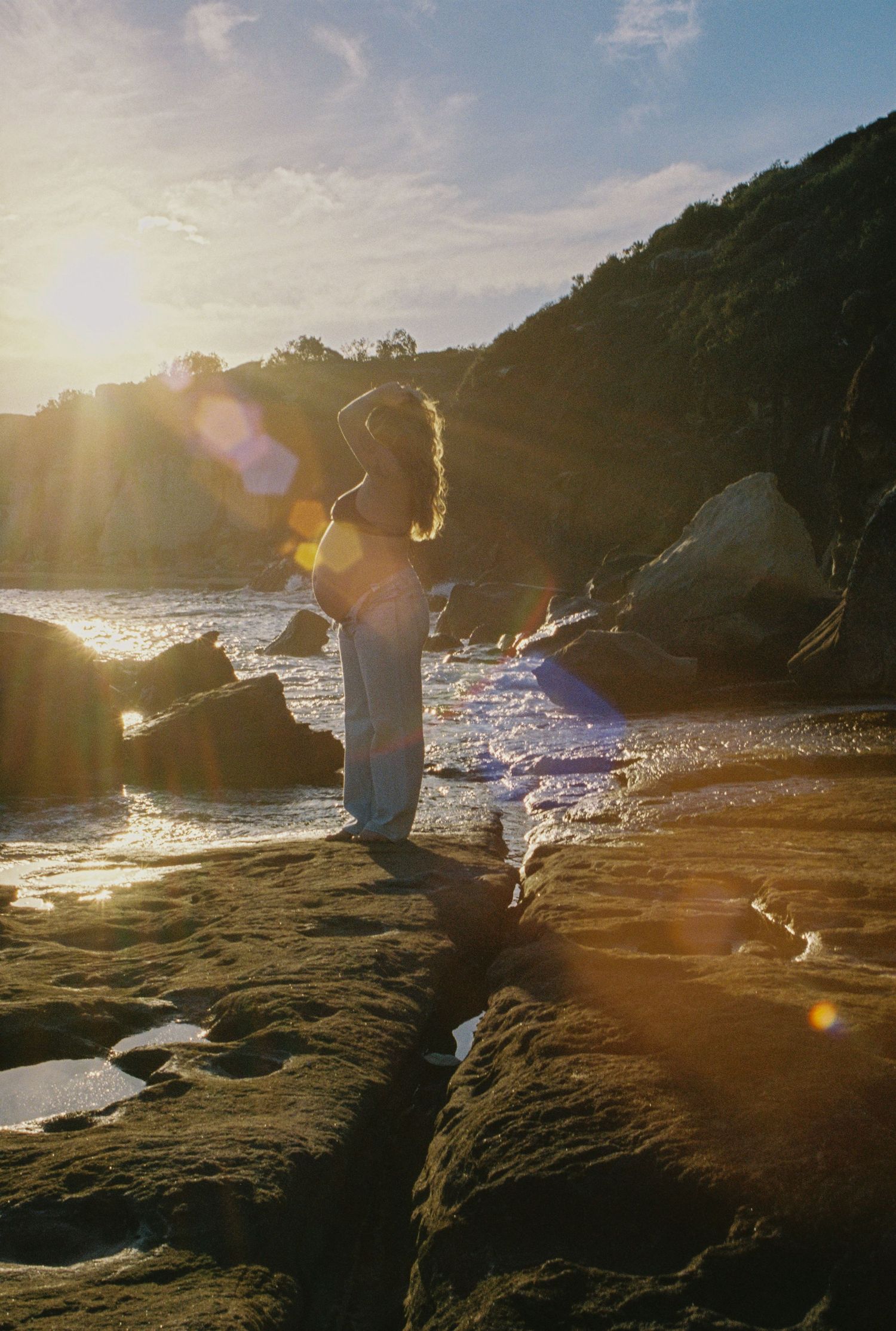 Woman standing on coastal rocks at golden sunset with ocean waves and lens flare creating a dreamy atmosphere.