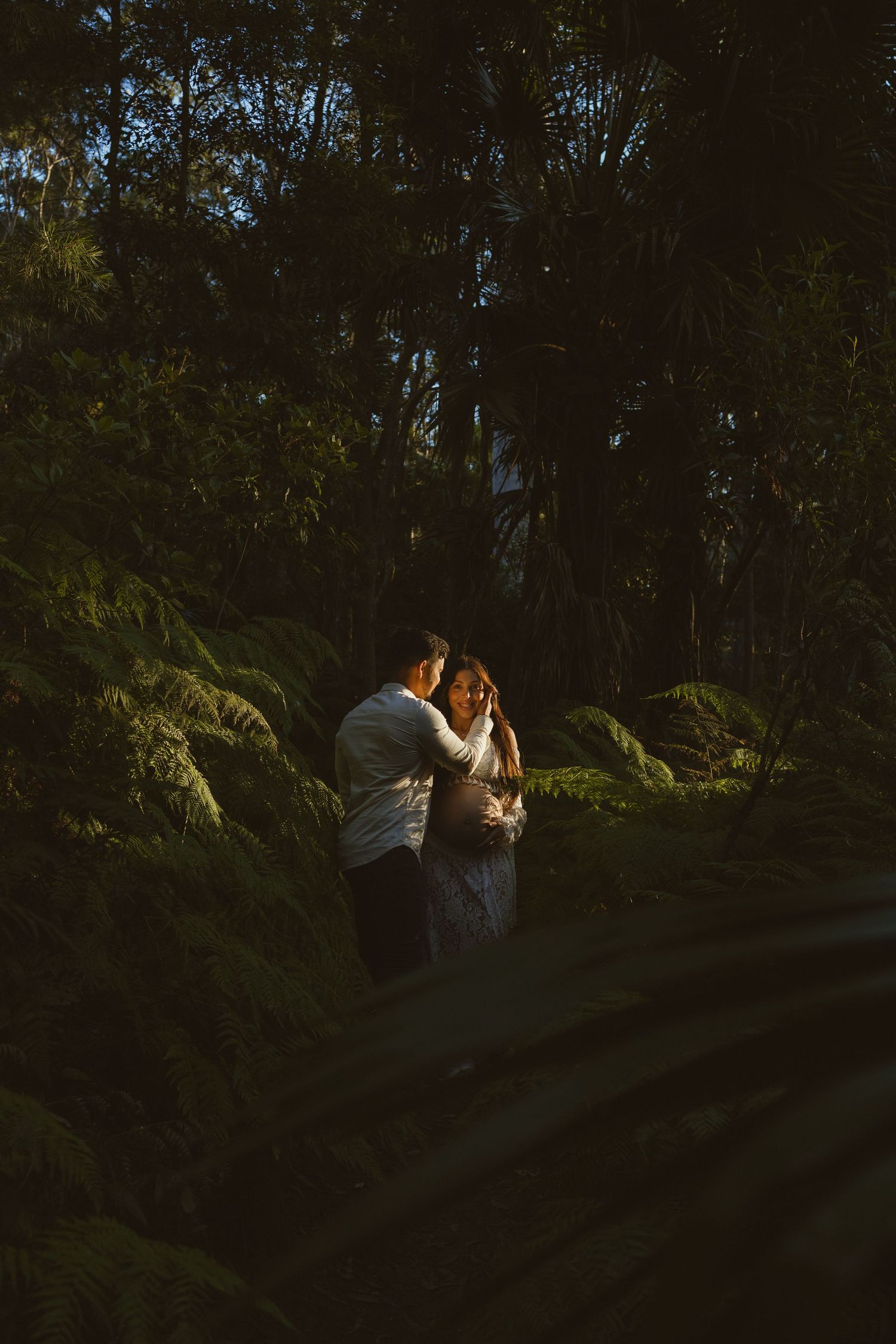 Couple embracing in a lush forest surrounded by ferns, bathed in warm golden light against dark foliage.
