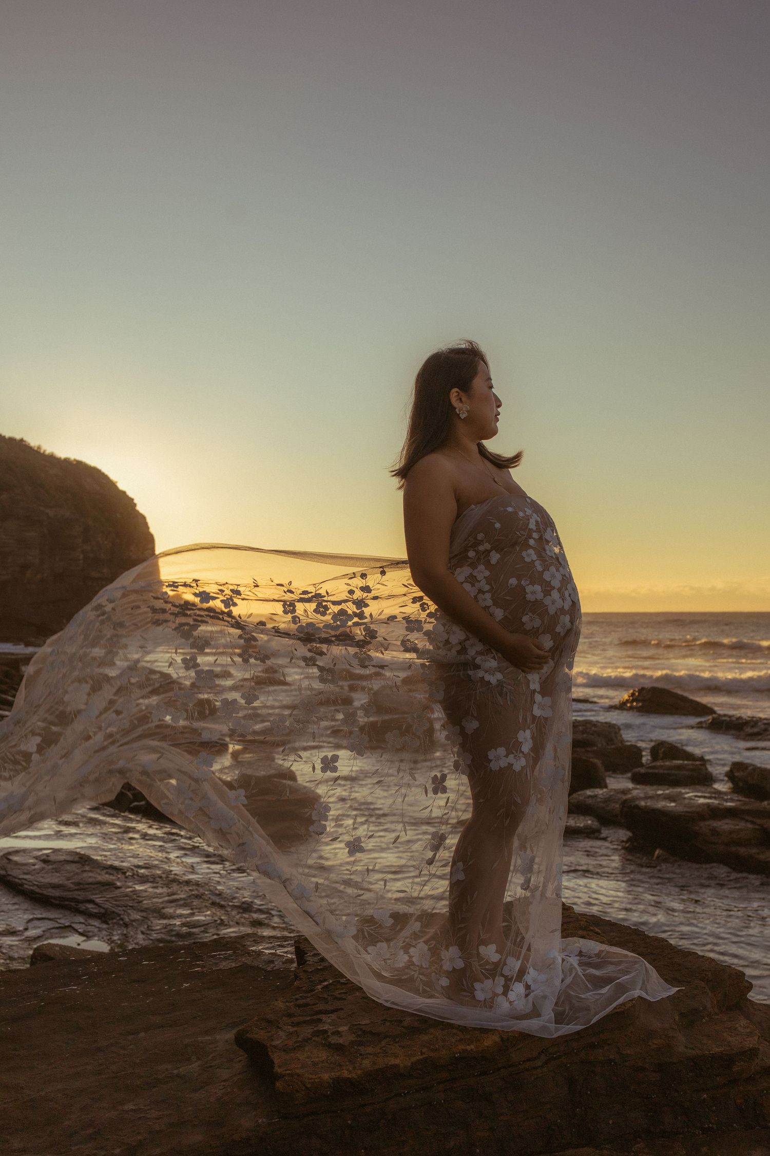 Pregnant woman in flowing lace gown stands on rocky beach at golden sunset, cradling her baby bump.