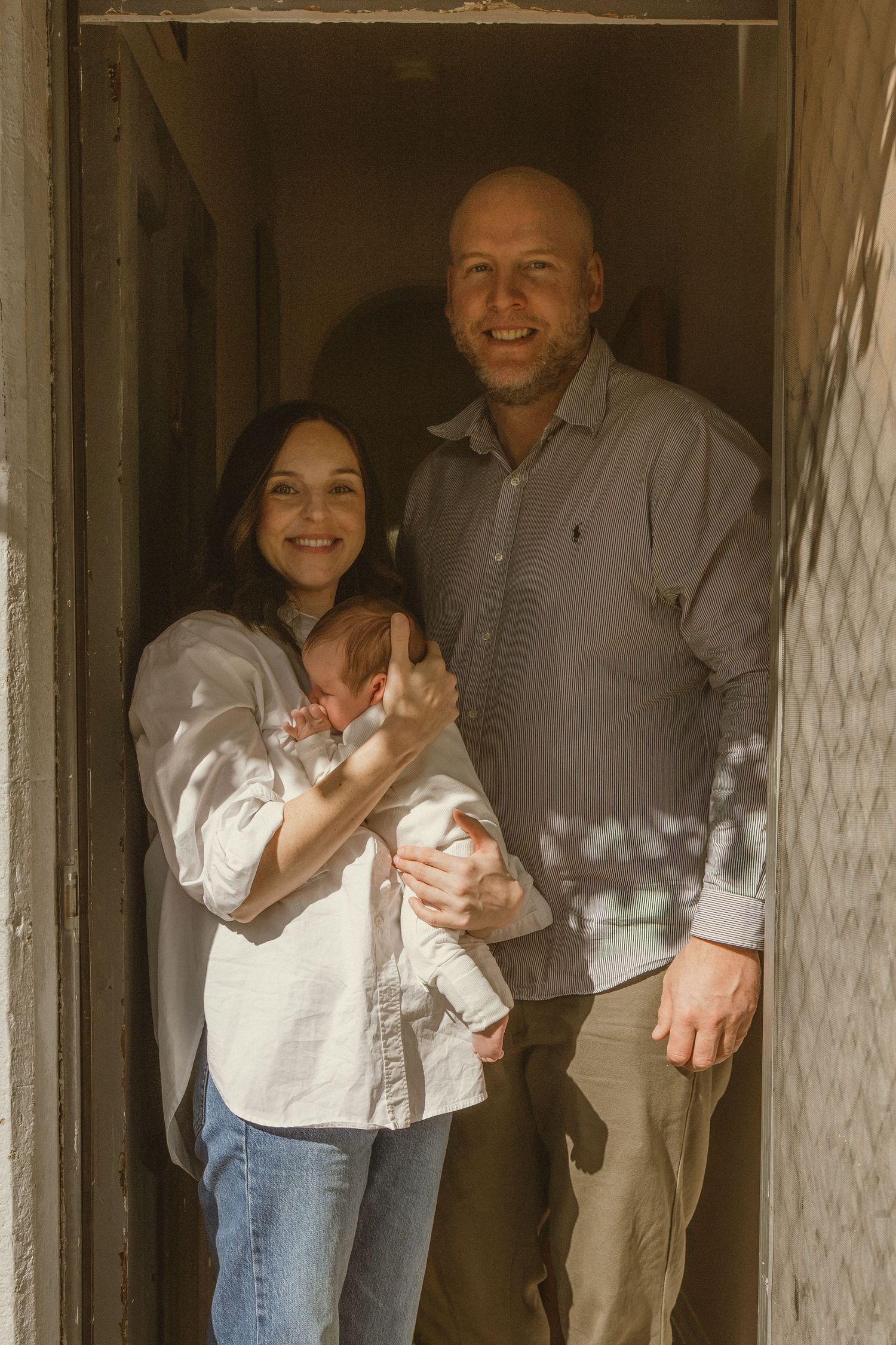 Happy couple standing in doorway holding their newborn baby, smiling warmly in natural sunlight.