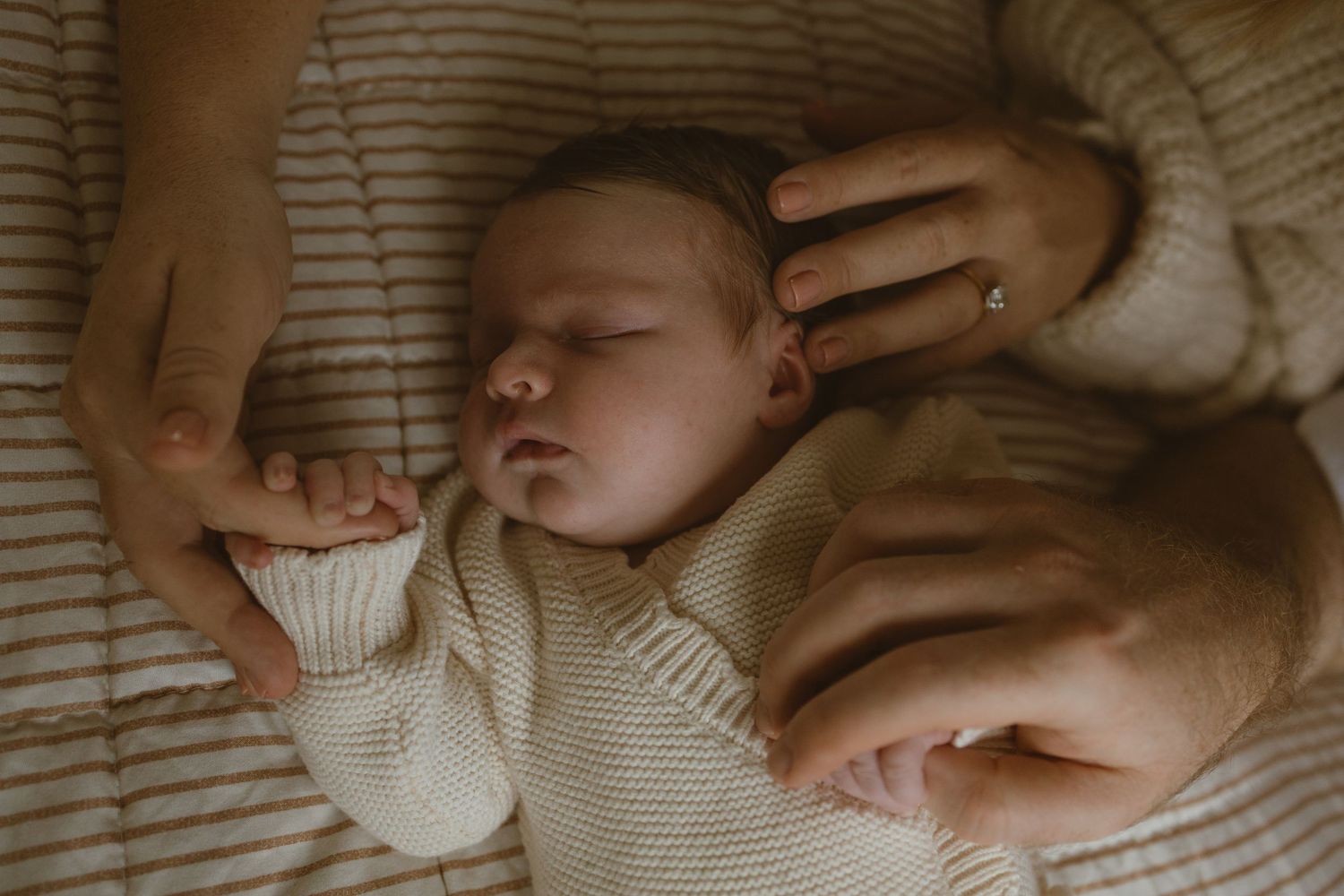 Sleeping newborn baby in cream knit onesie held gently by mother's hands on striped blanket.