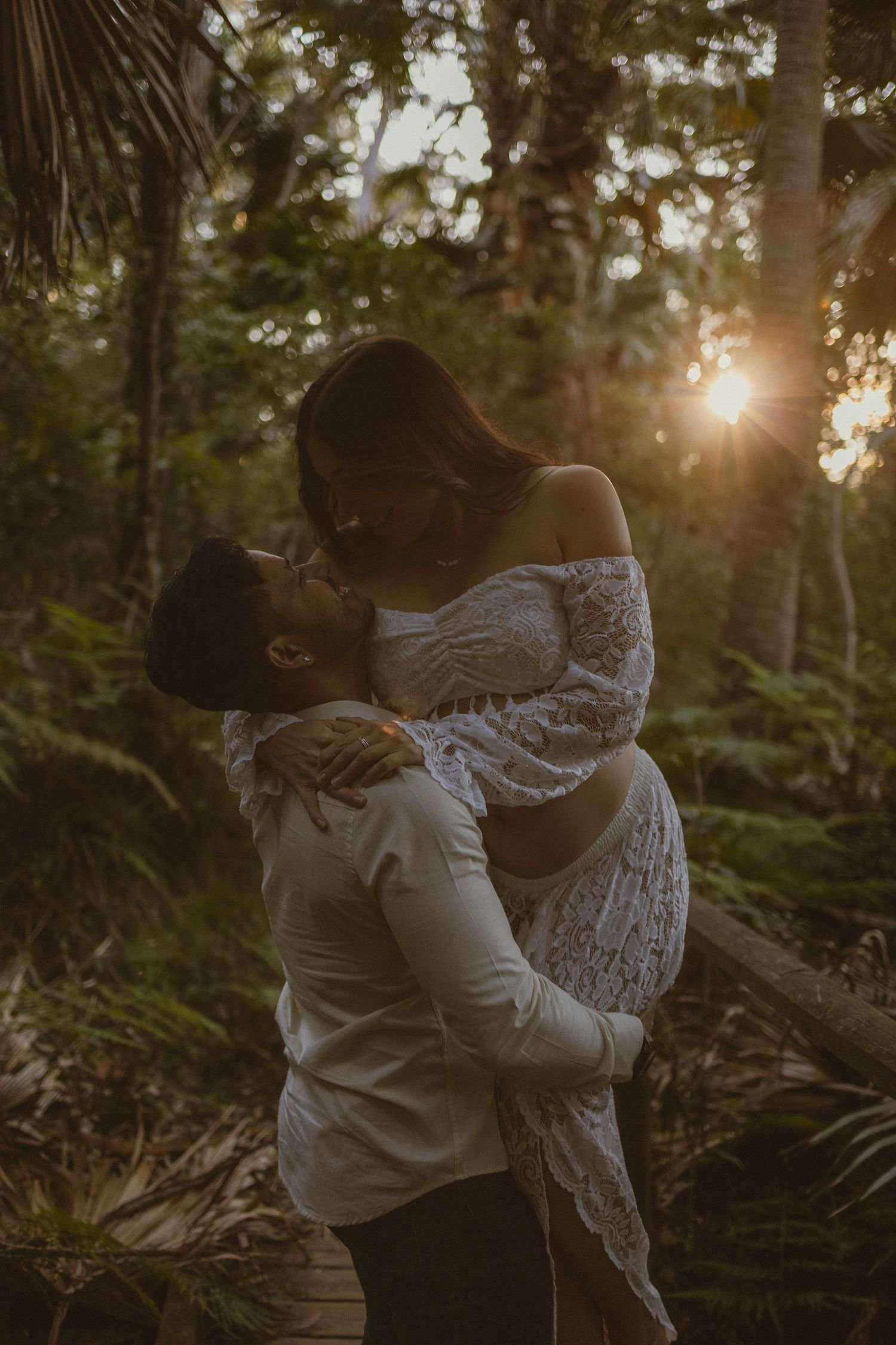 Couple sharing romantic kiss in sunlit tropical forest, pregnant woman in white lace dress lifted by partner.