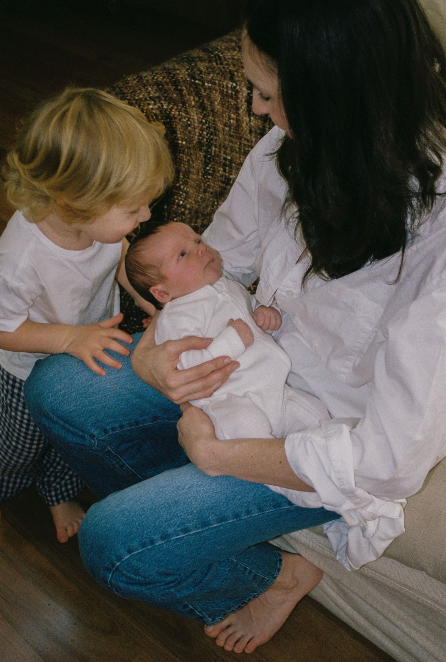 A mother in a white shirt and jeans sits on the floor holding a newborn while a toddler looks on lovingly.