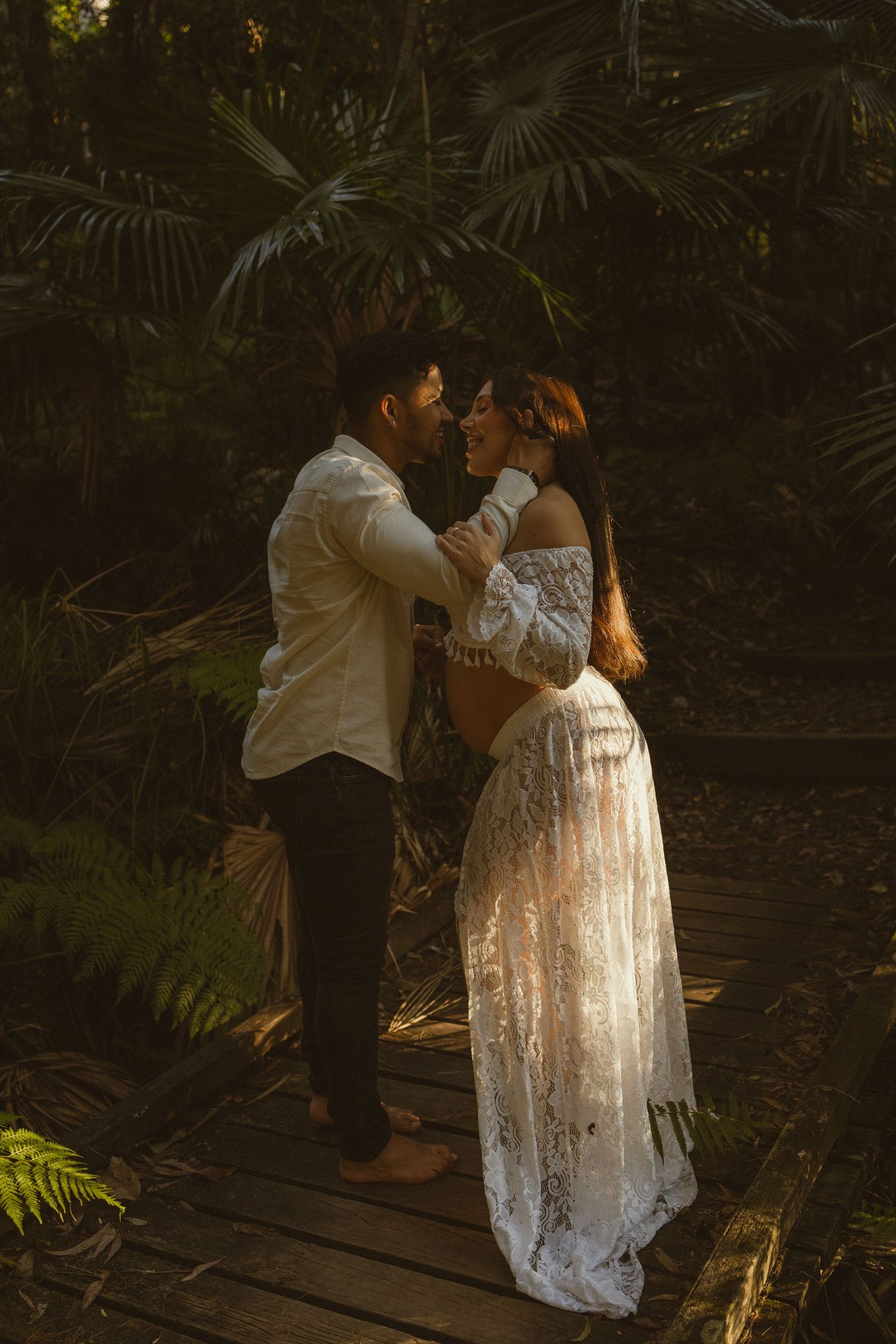 Couple sharing a romantic moment in a lush tropical garden, woman in white lace off-shoulder gown.