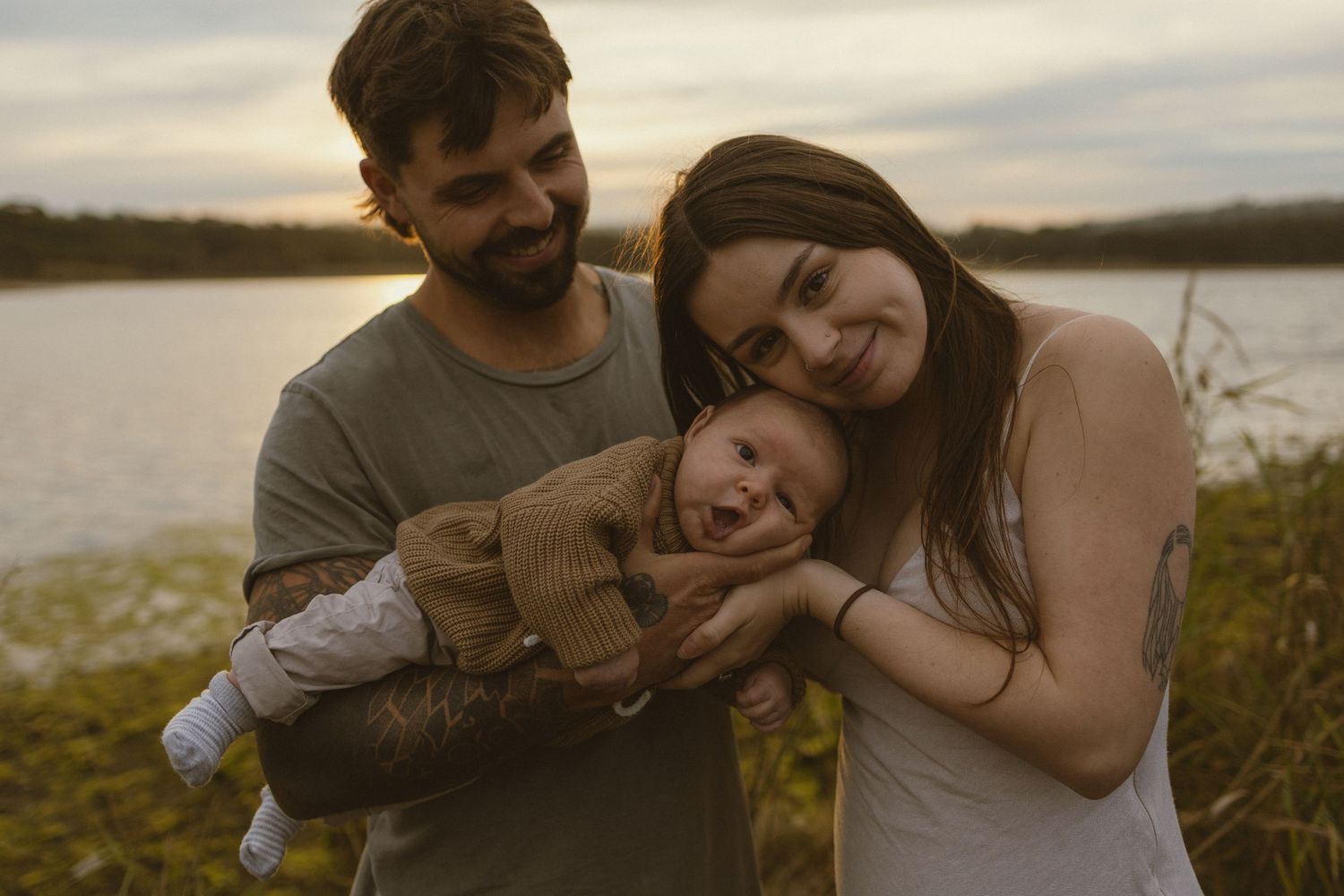 Young couple smiling and holding their laughing newborn baby outdoors near a lake at sunset.