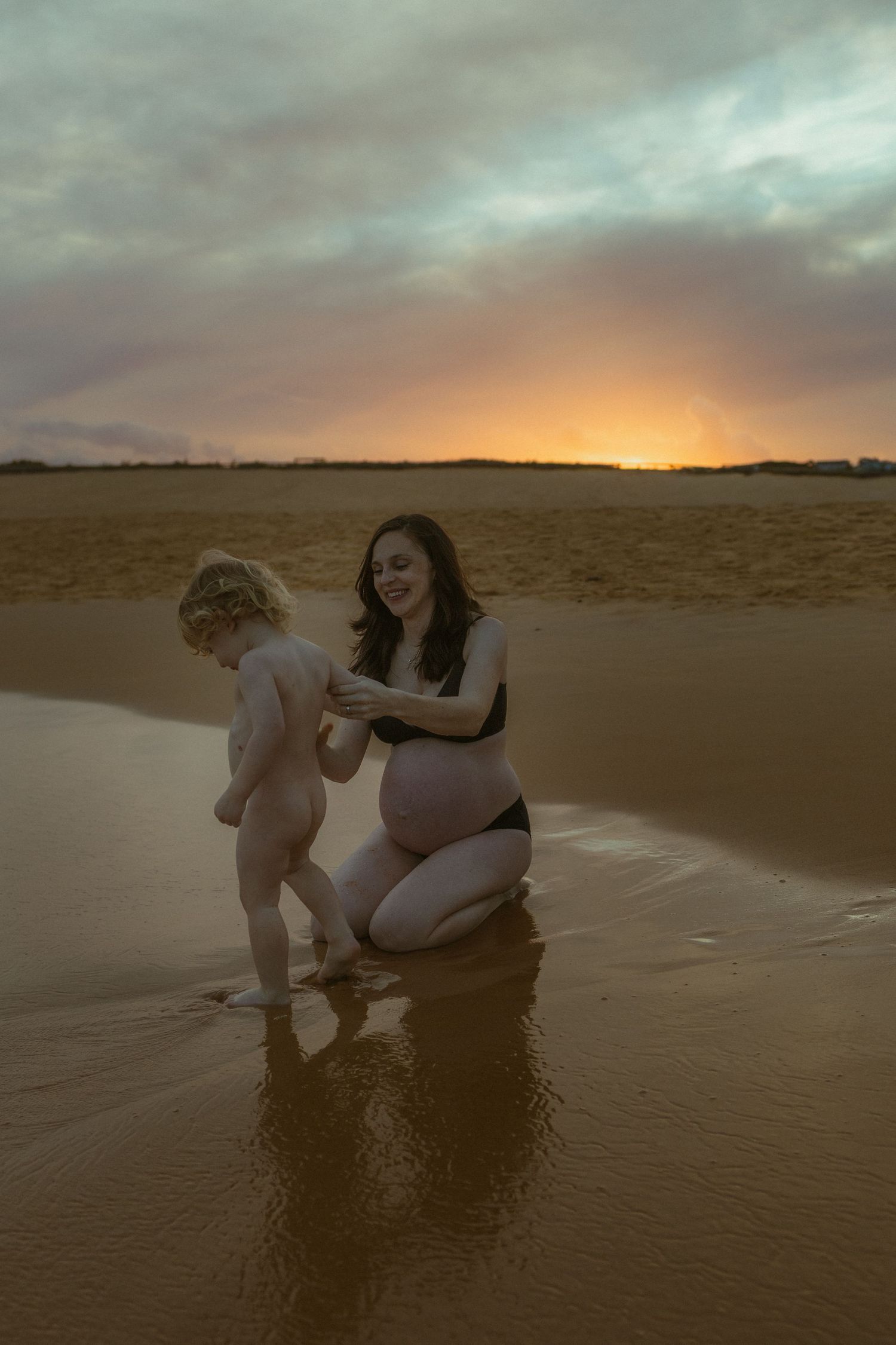 Pregnant woman in black swimsuit kneels on sandy beach with toddler at sunset, shallow water reflecting warm orange sky.