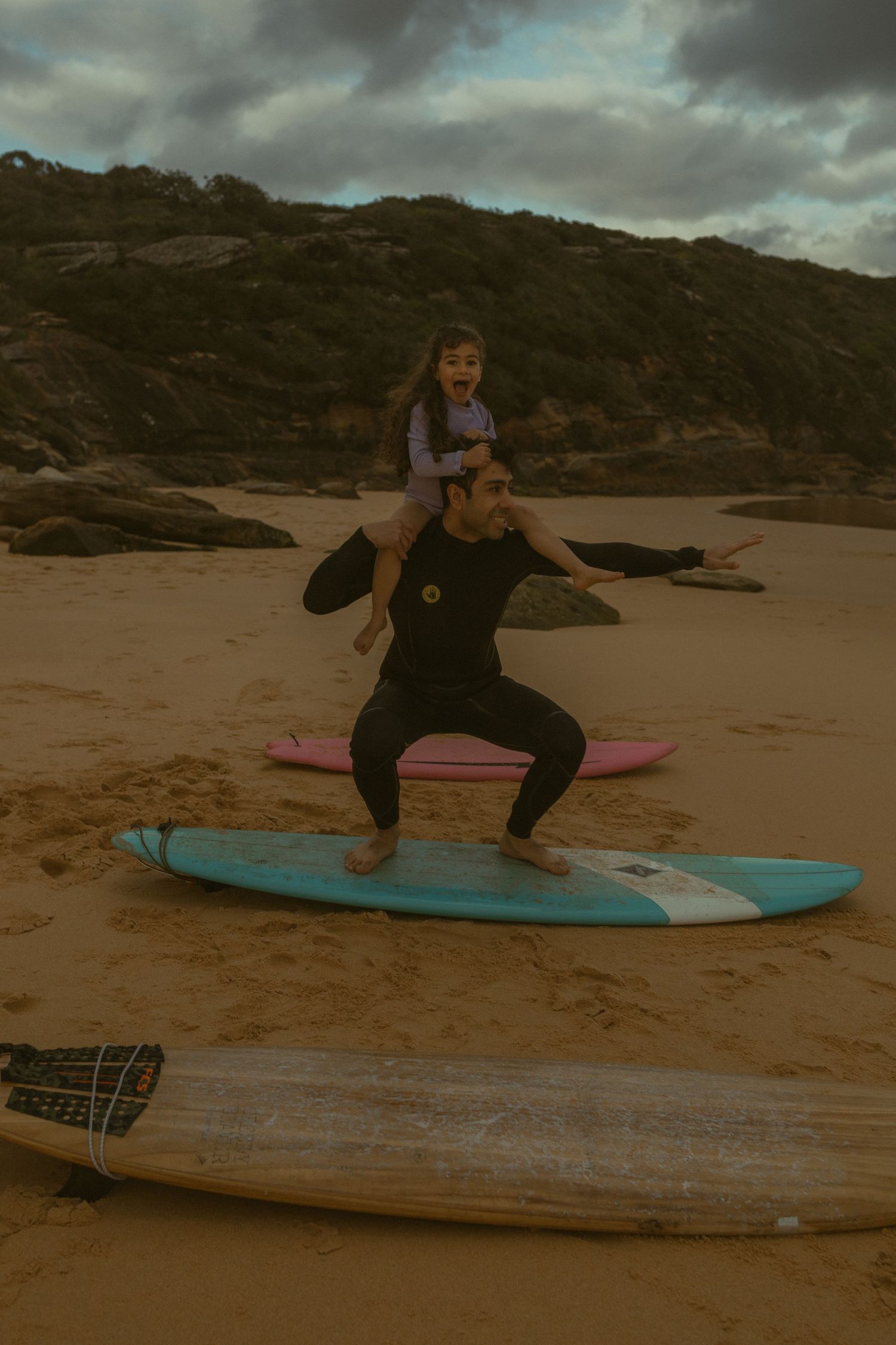 Man in wetsuit practices surfing stance on beach with child on shoulders, blue surfboard beneath them on sandy shore.