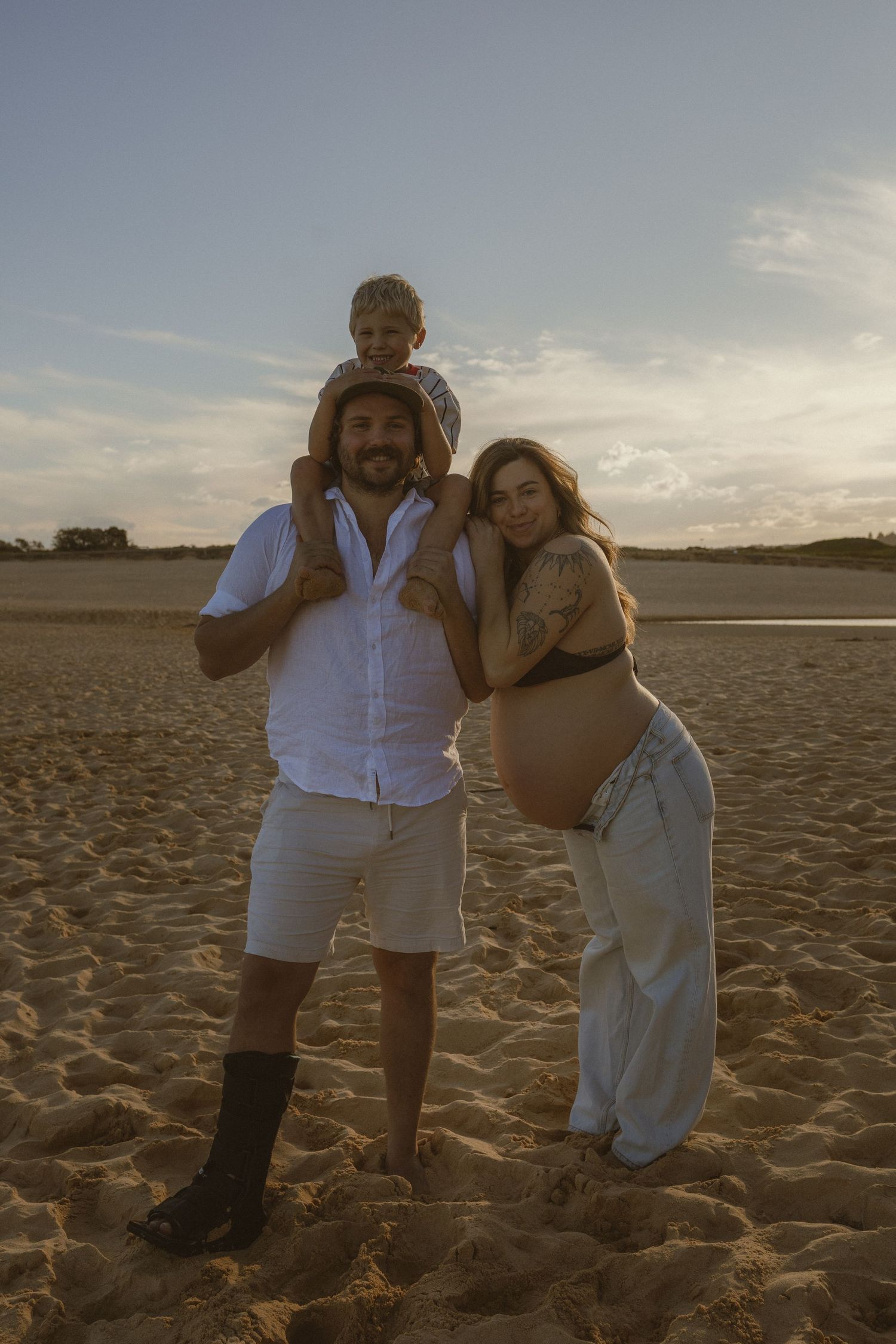 A pregnant woman and man with child on shoulders pose on sandy beach at sunset.