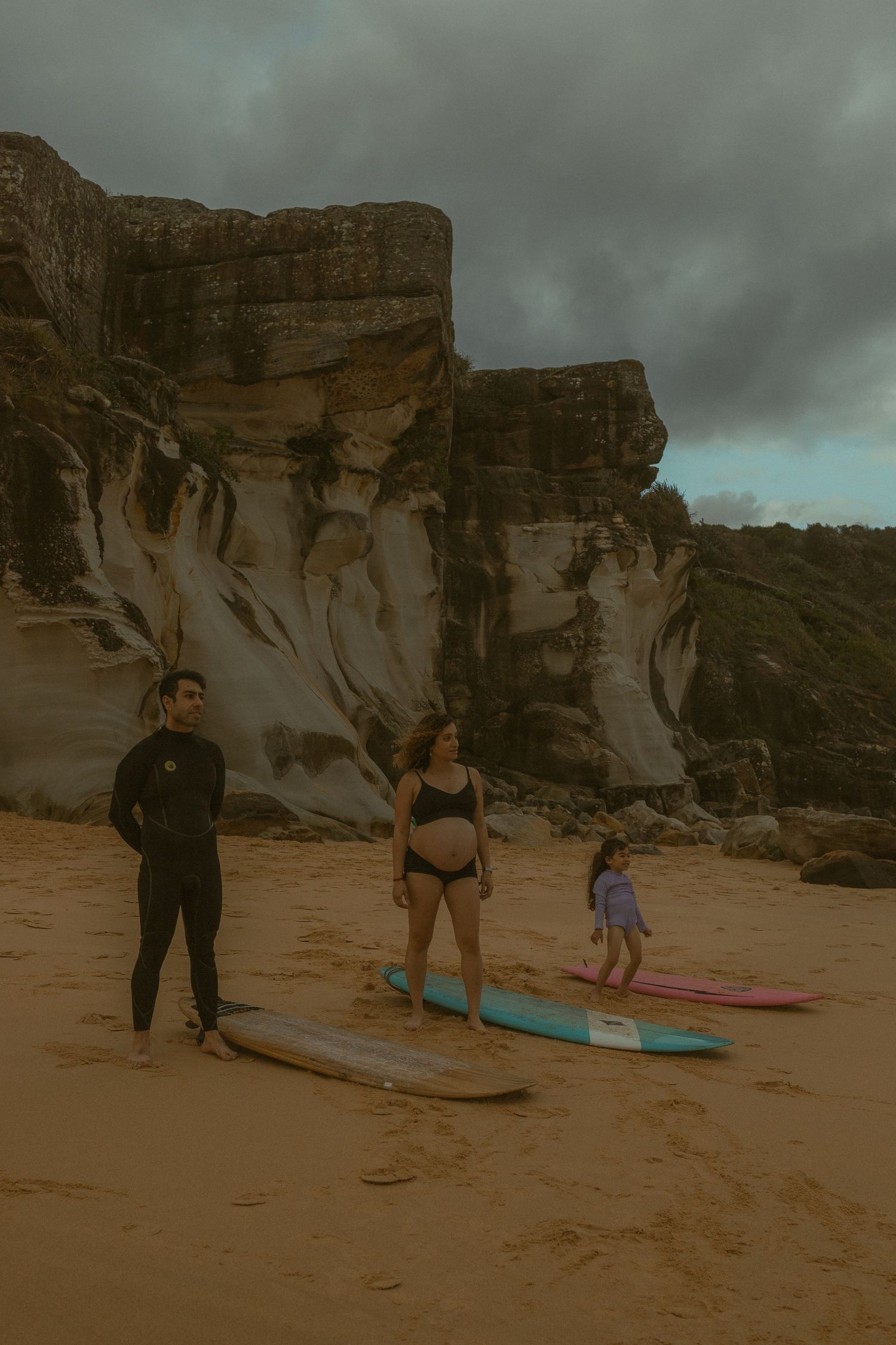 Family with surfboards on sandy beach near dramatic sandstone cliffs under overcast sky.
