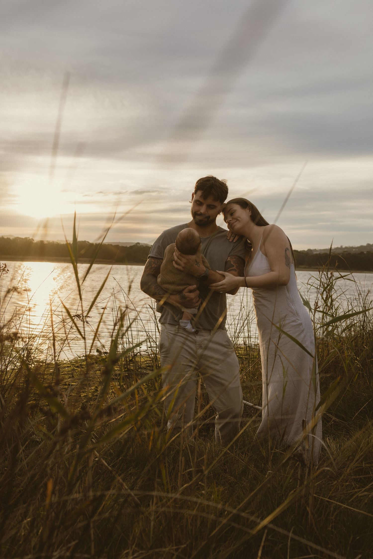 A couple stands in tall grass by a lake at sunset, tenderly gazing at their newborn baby in a golden evening light.