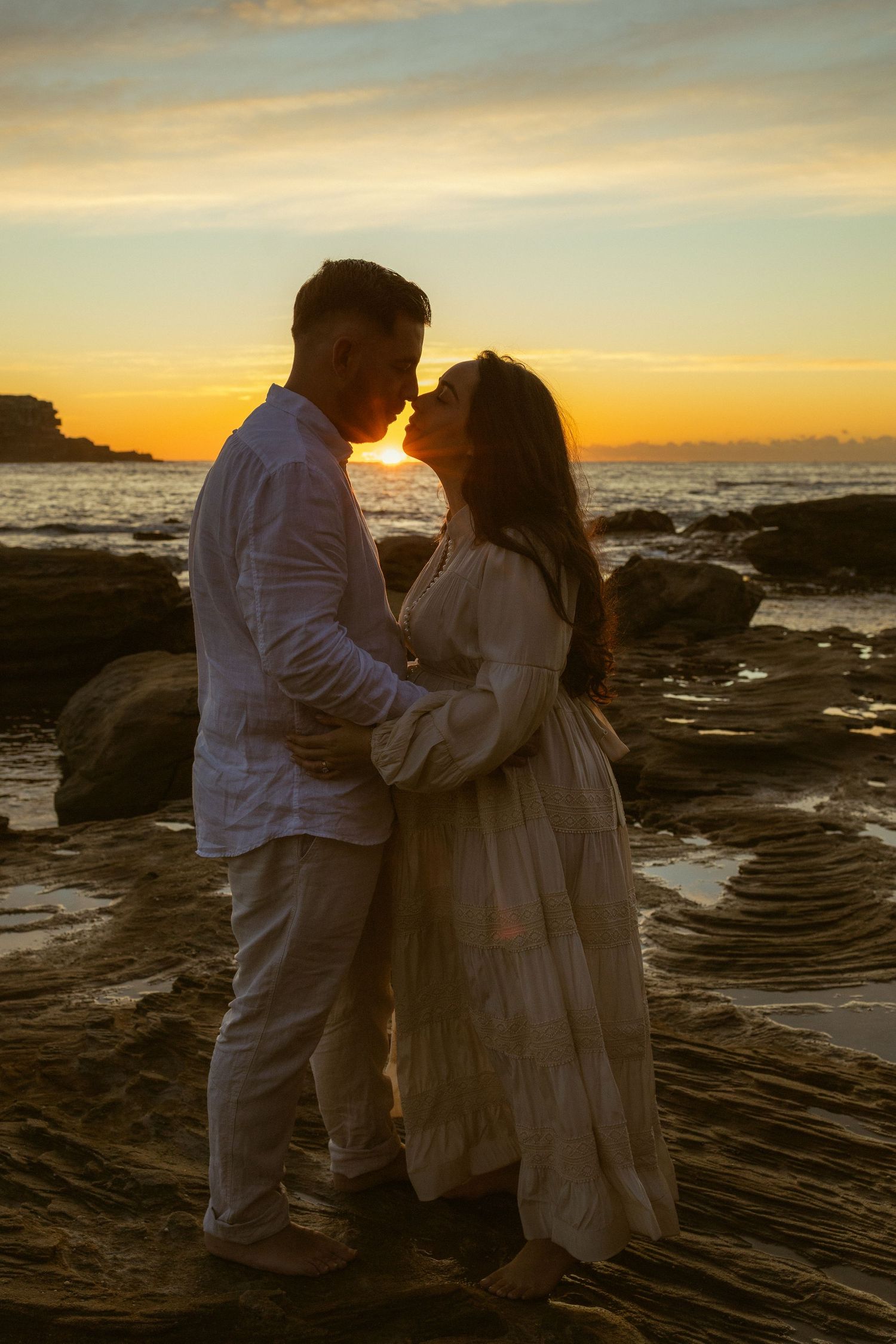 Romantic couple in white embracing on rocky beach at golden sunset with ocean backdrop.