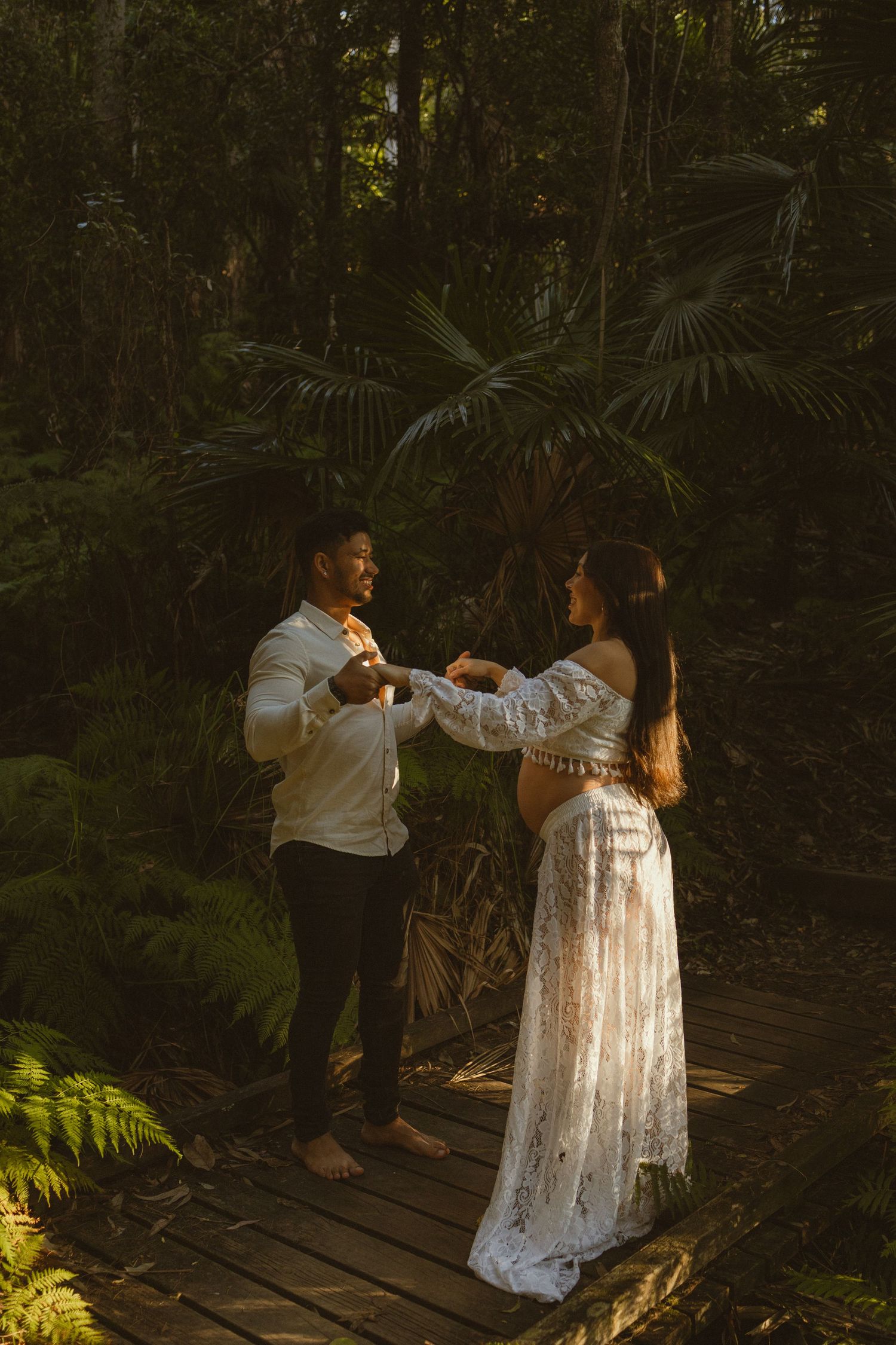 Couple holding hands on a wooden deck surrounded by lush tropical forest during a golden hour maternity photoshoot.