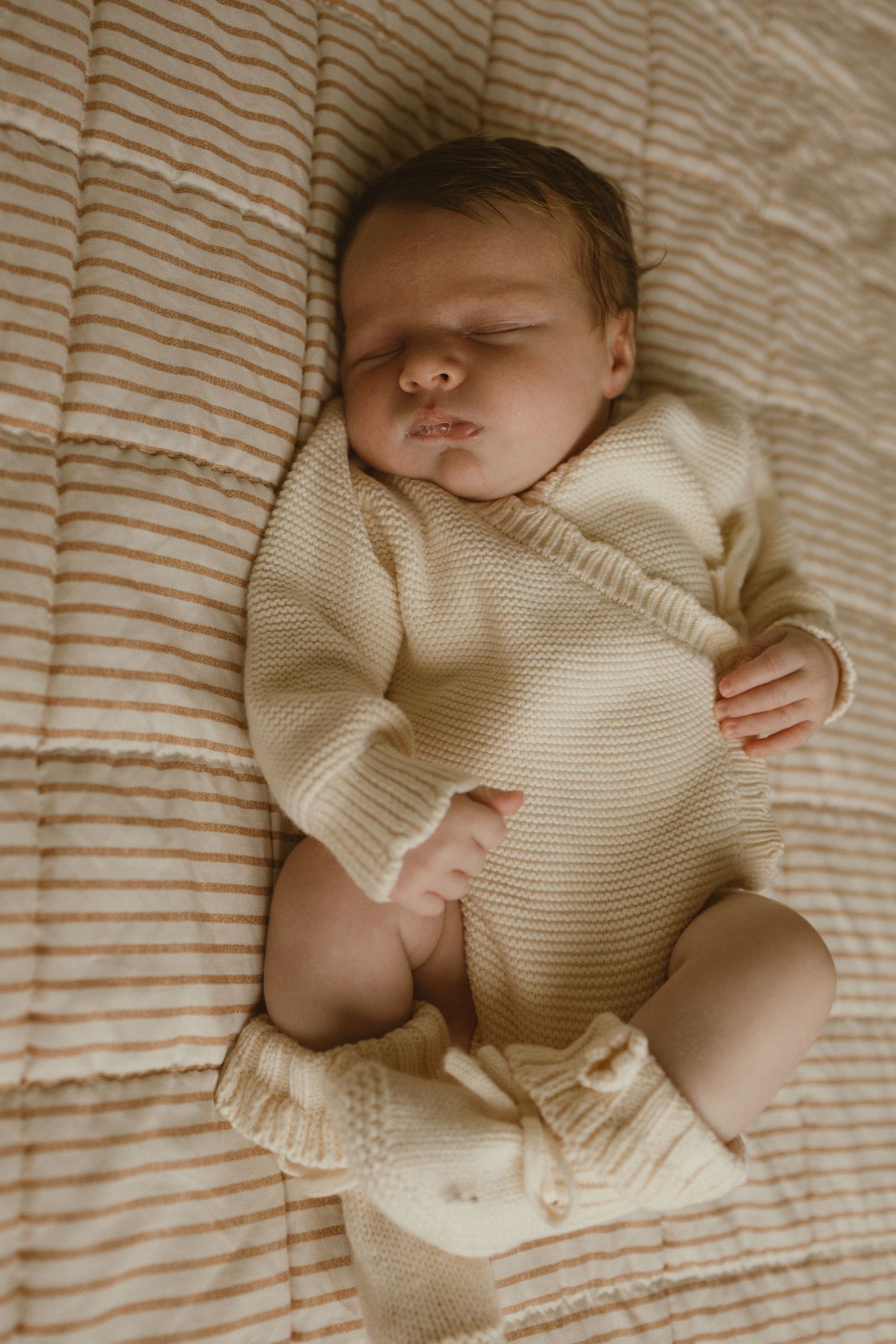 Sleeping newborn baby in cream knit romper lying on striped beige blanket, peaceful infant portrait photography.