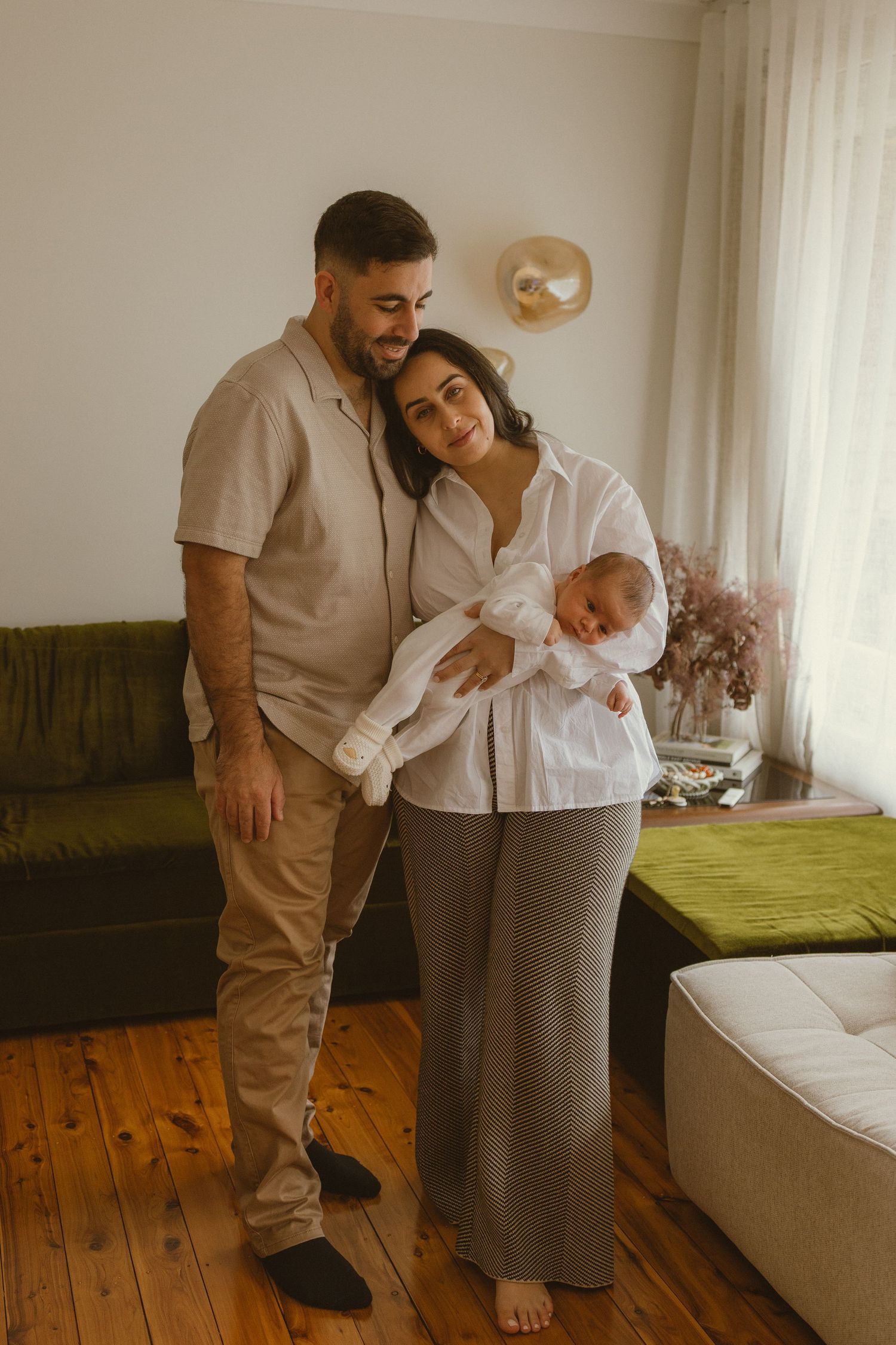 A couple stands together in a cozy living room, the woman cradling a newborn baby in her arms.