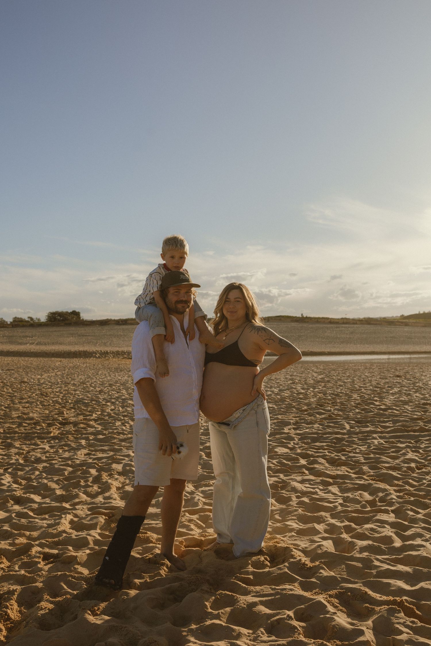Pregnant woman poses with partner and child on sandy beach at golden hour sunset.