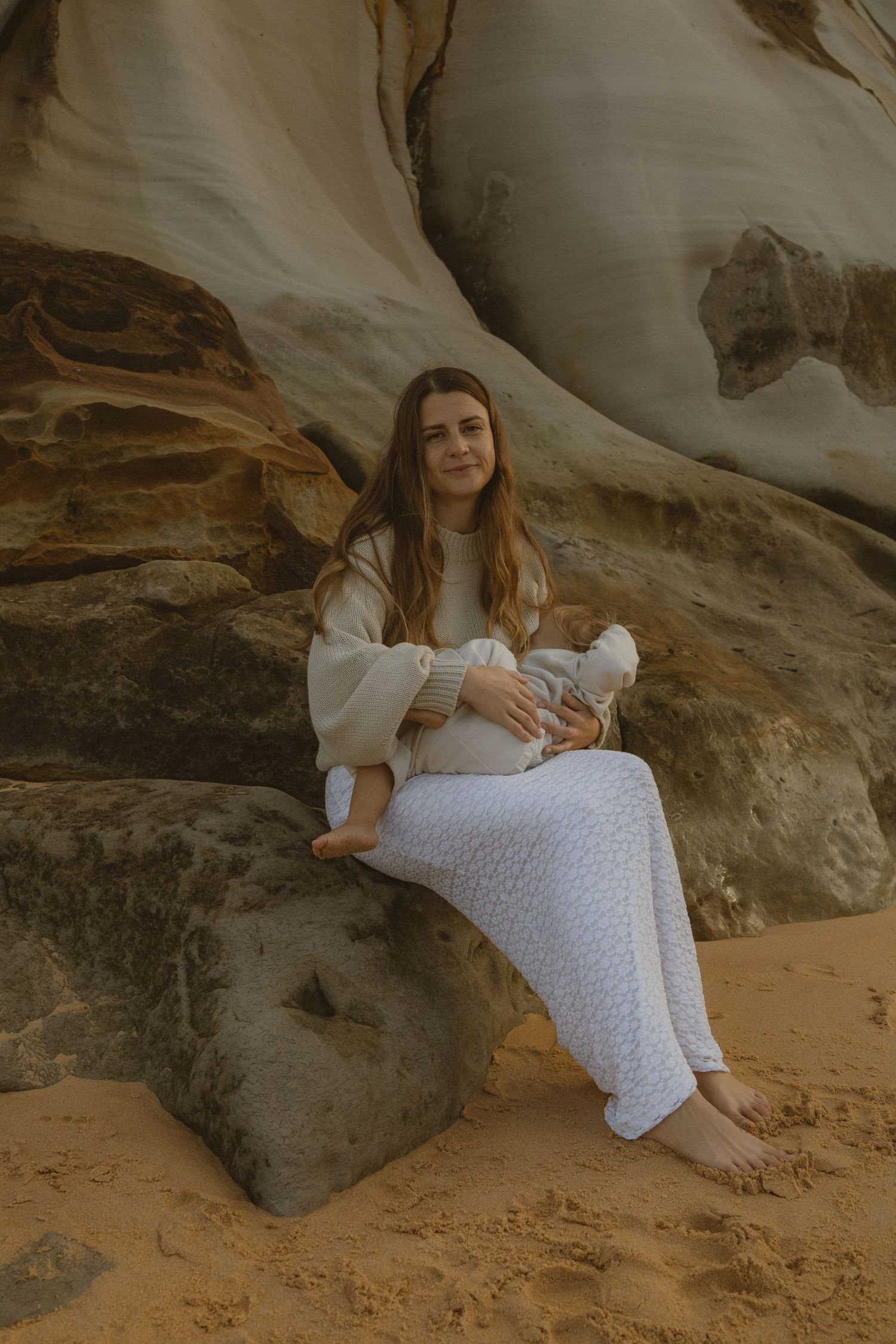 A barefoot woman in white skirt sits on sandstone rocks cradling her baby in a serene coastal setting.