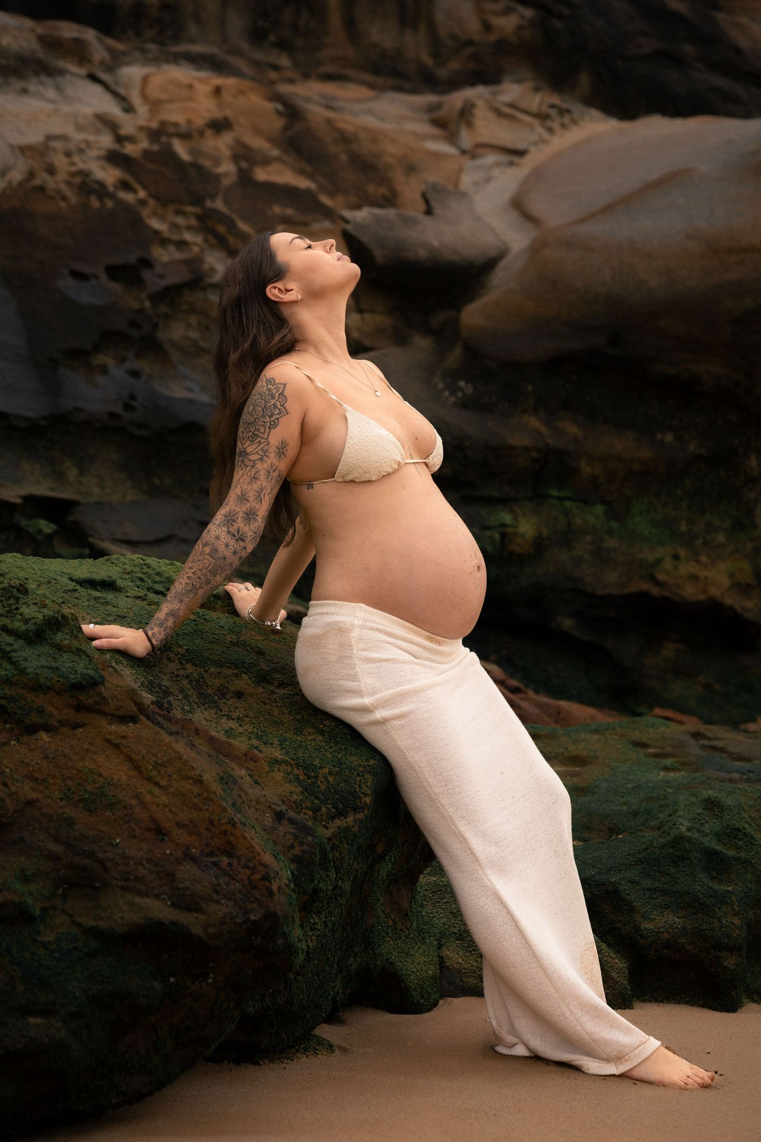 A pregnant woman in white dress poses gracefully against coastal rock formations at sunset.