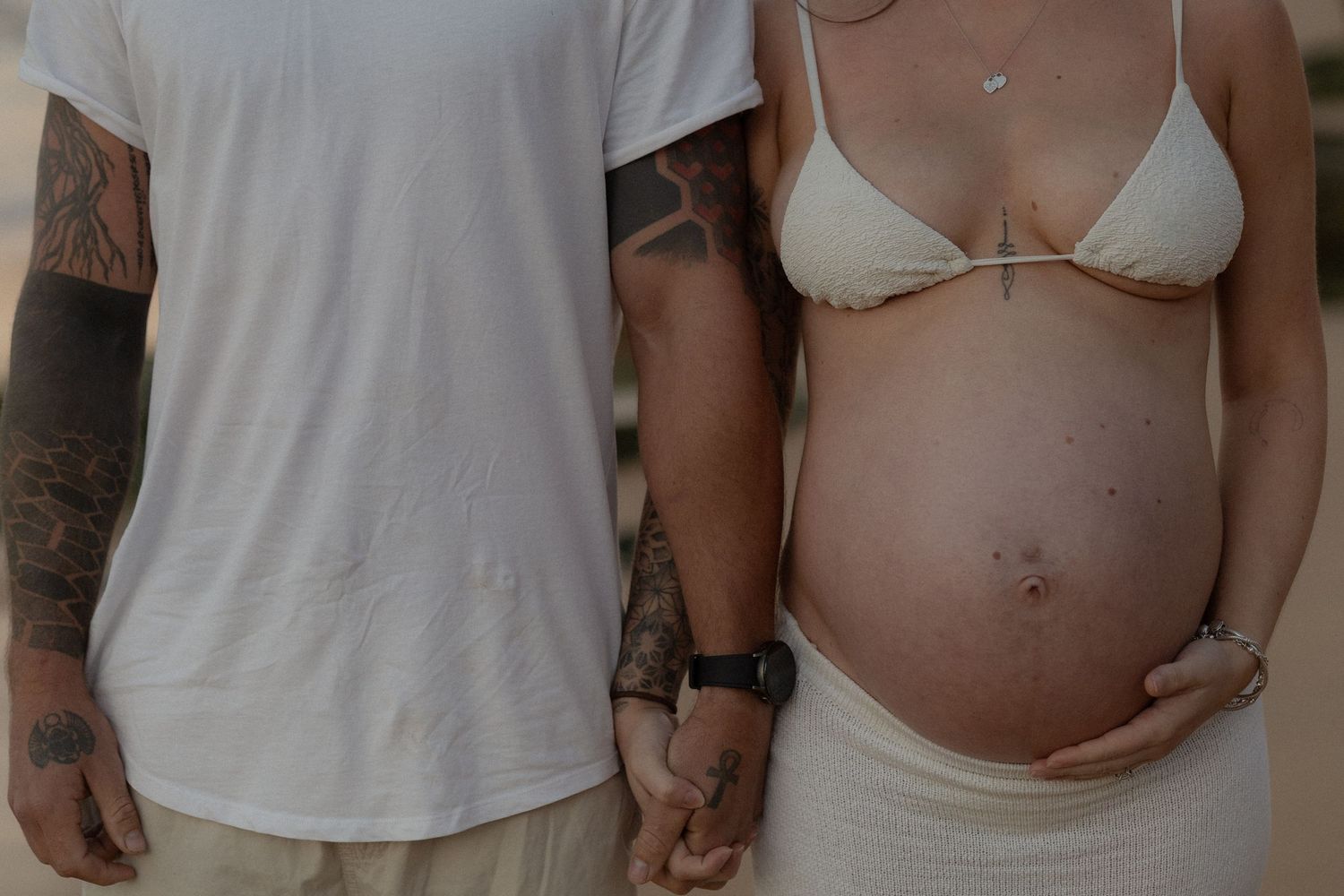 Pregnant woman in white bikini holding baby bump beside tattooed man in white t-shirt on beach.