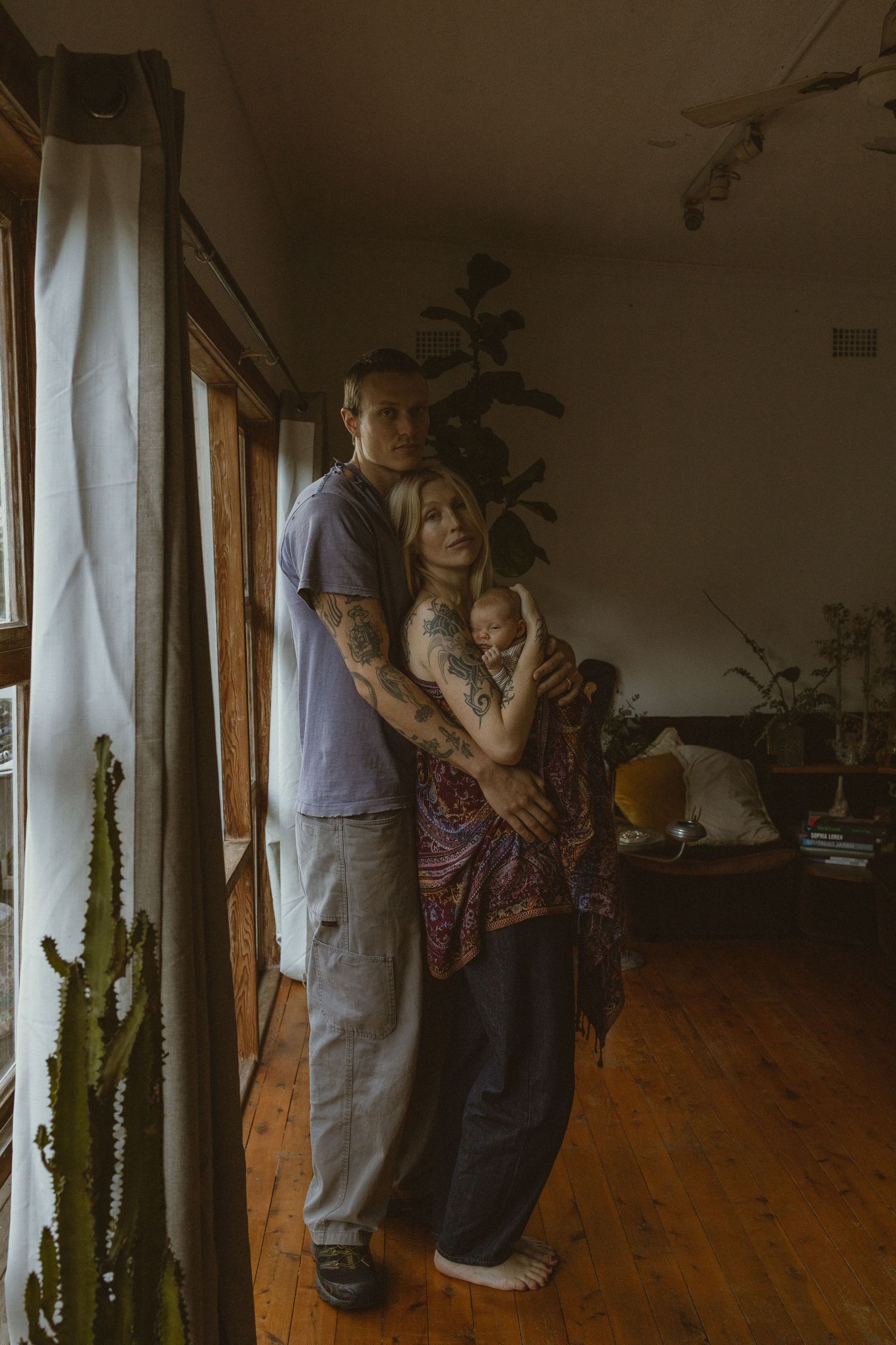 Tattooed couple holding newborn baby by window in rustic living room with hardwood floors and indoor plants.