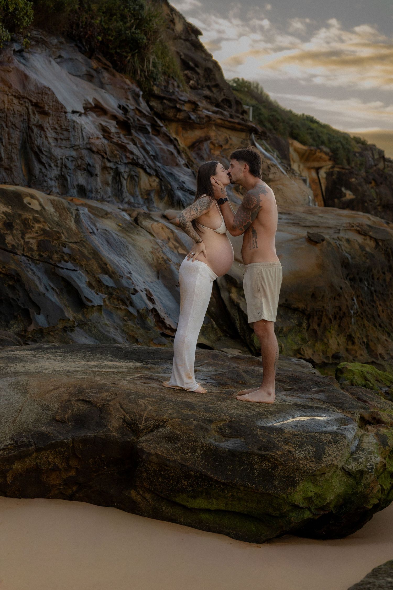 Pregnant couple sharing a kiss on coastal rocks at sunset, woman in white skirt, man shirtless in beige shorts.