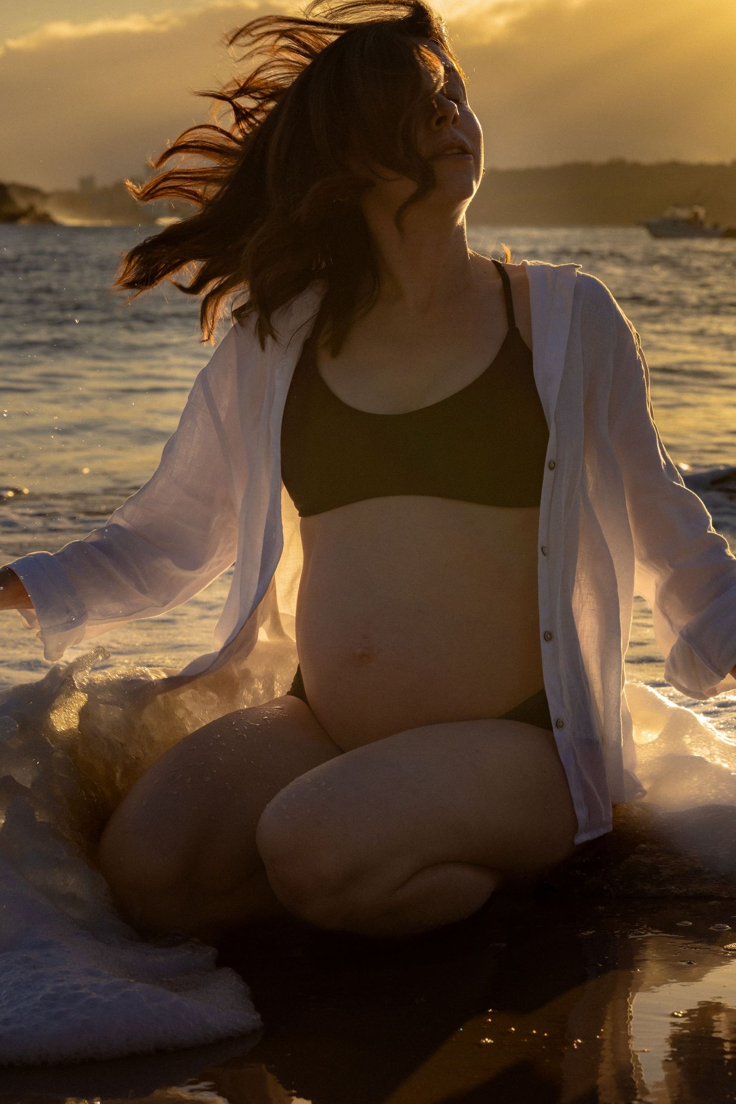 Pregnant woman in bikini top and white shirt sits on beach at golden sunset with hair blowing in wind.