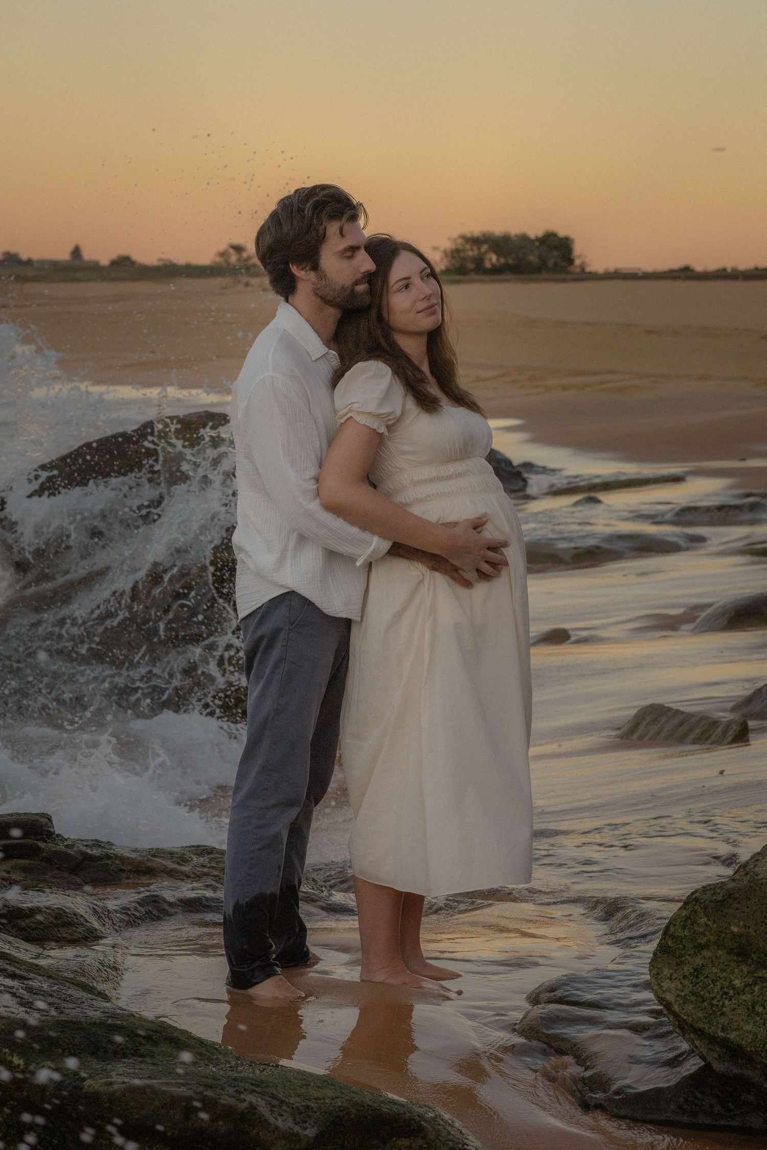 Couple embracing at sunset on rocky beach, pregnant woman in white dress, waves crashing behind them.