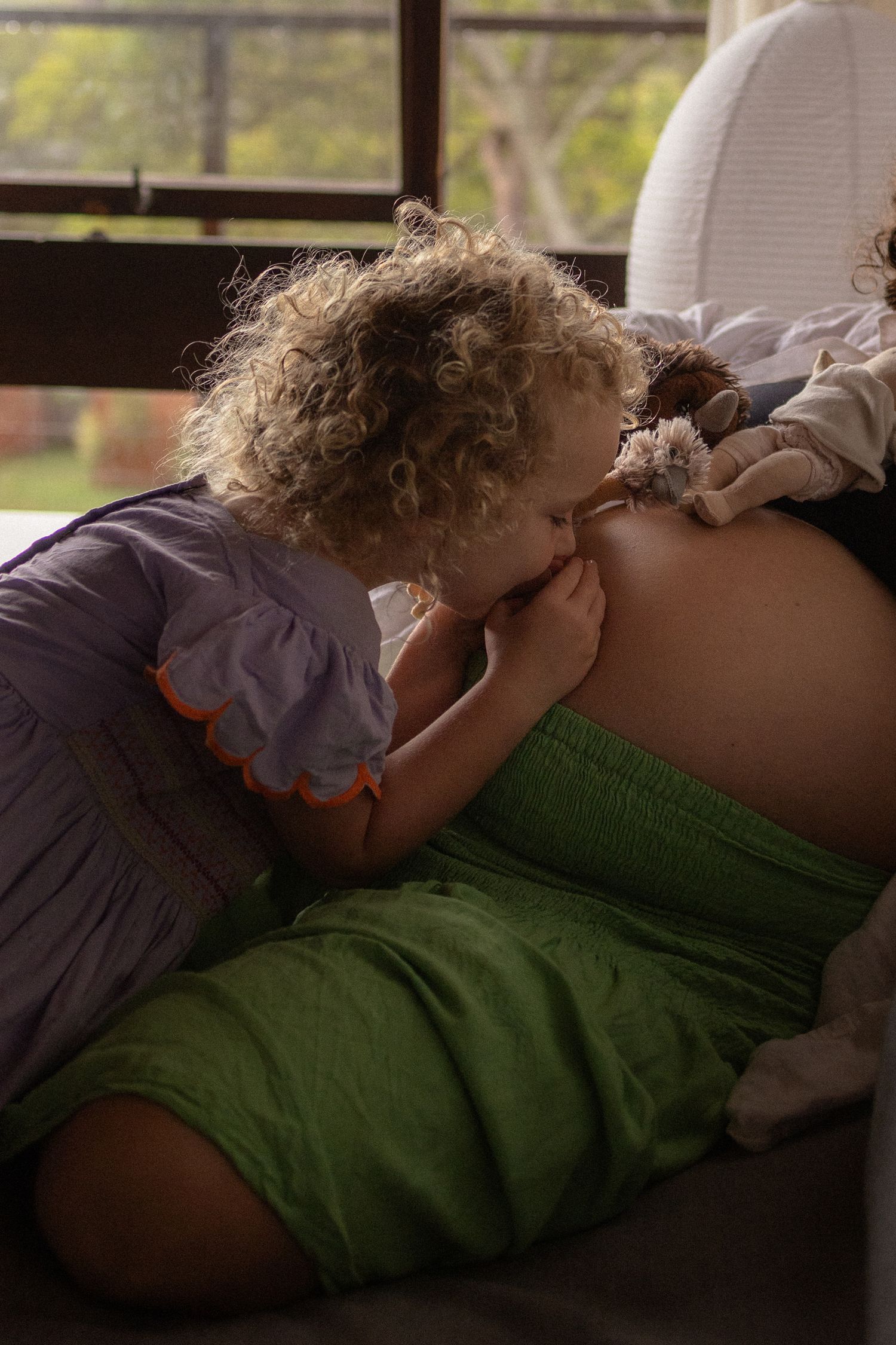 Young curly-haired child kissing pregnant mother's belly while holding a stuffed toy indoors.