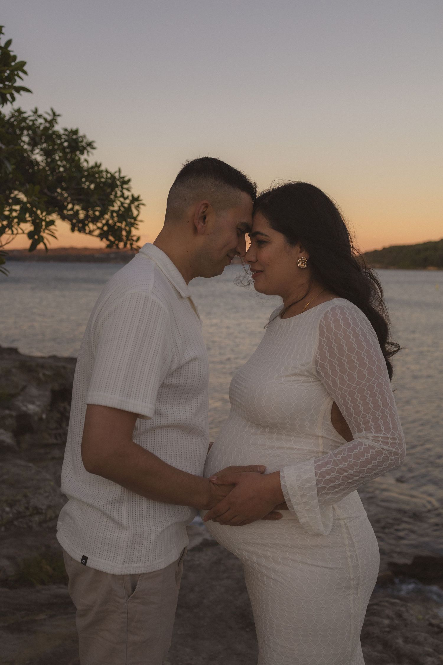 Pregnant couple in white outfits touching foreheads tenderly by a sunset beach, cradling baby bump.