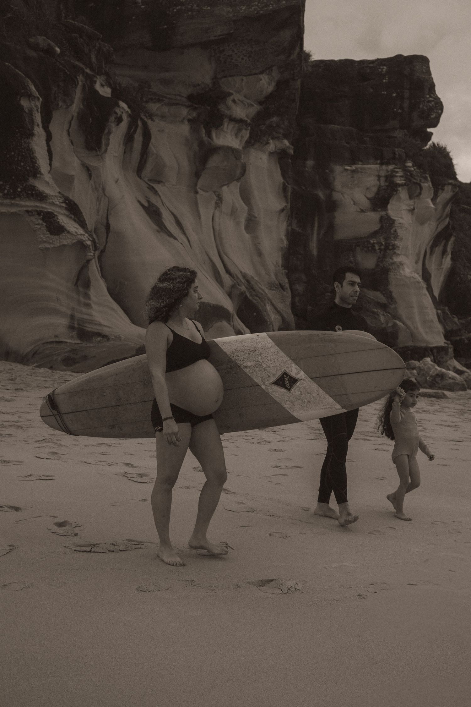 Pregnant woman in bikini carrying surfboard on sandy beach with man and child, rocky cliffs in sepia background.