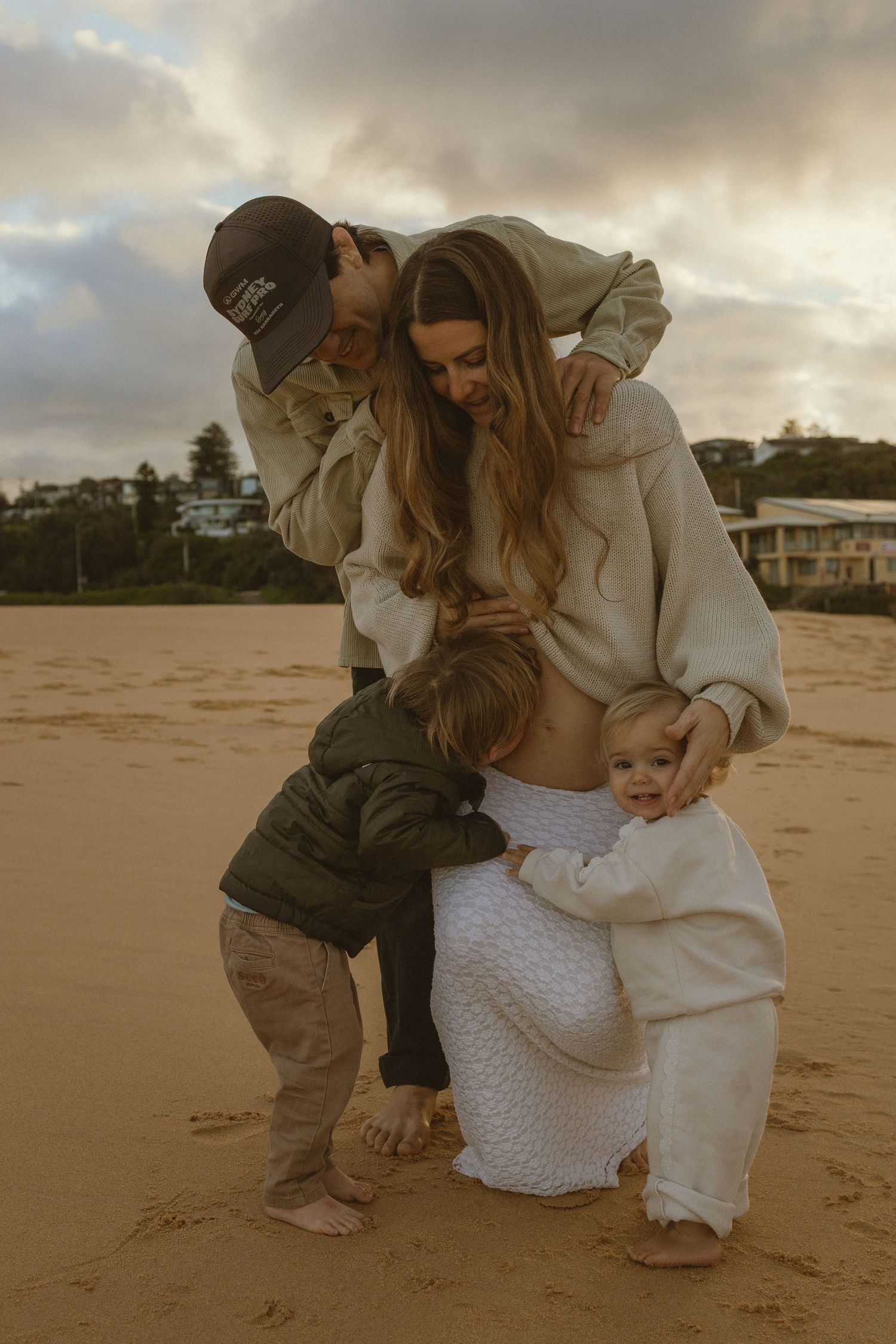 Family of four sharing a tender moment on a sandy beach at sunset, with two young children hugging their parents.