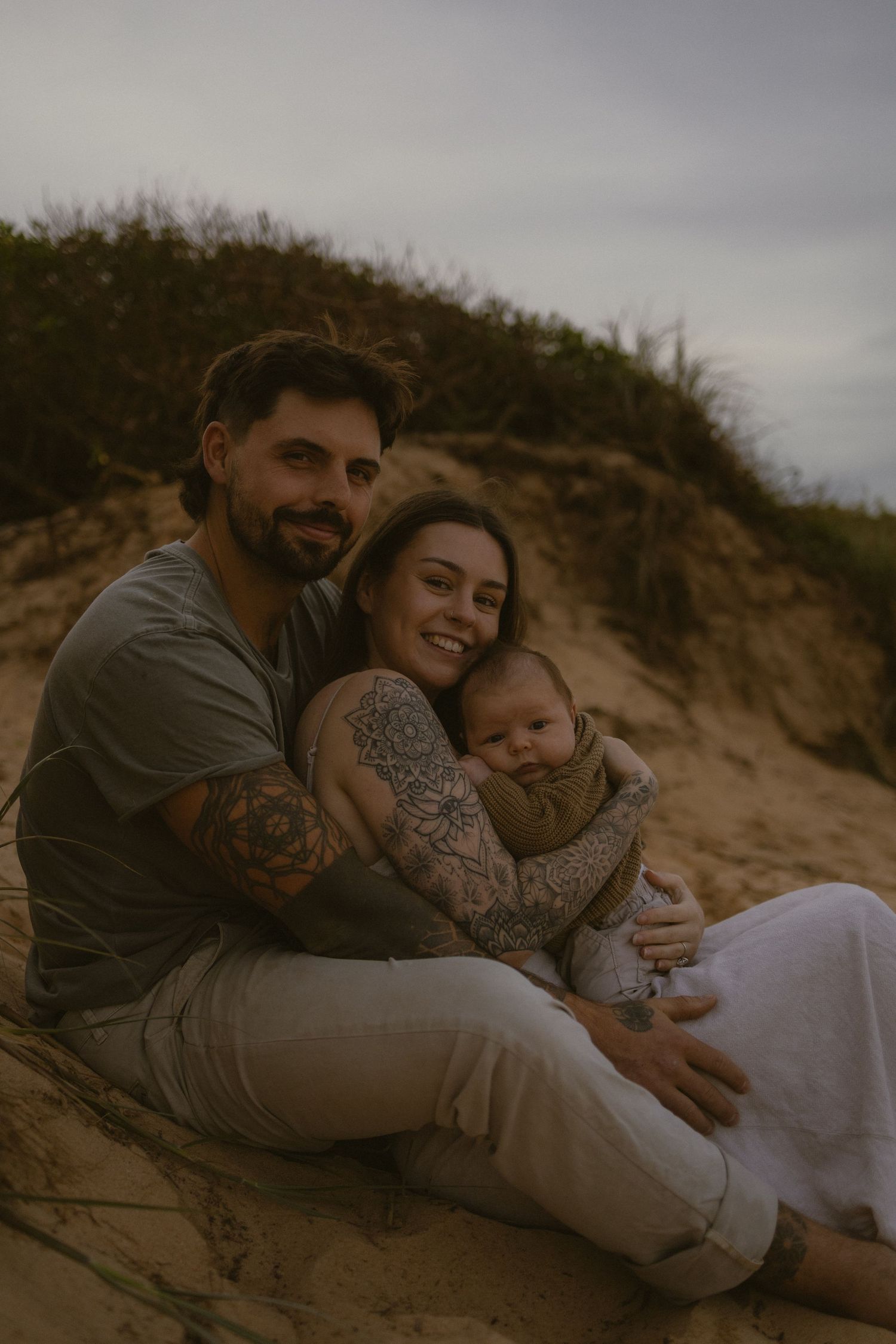 A tattooed couple sits on sandy beach dunes, smiling while holding their newborn baby in a cozy family portrait.