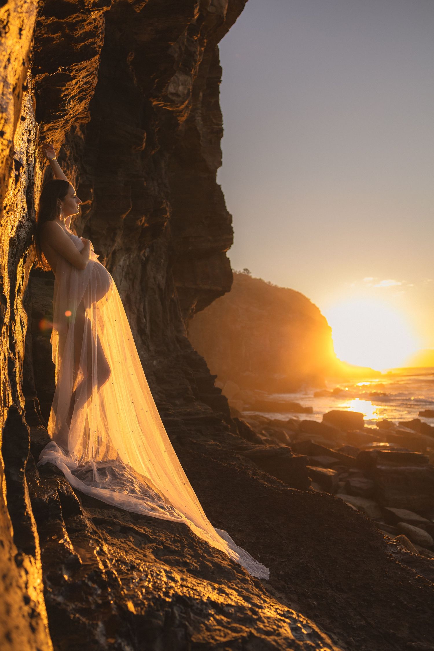 Silhouette of pregnant woman in sheer white dress framed by rocky cliffs at sunset.