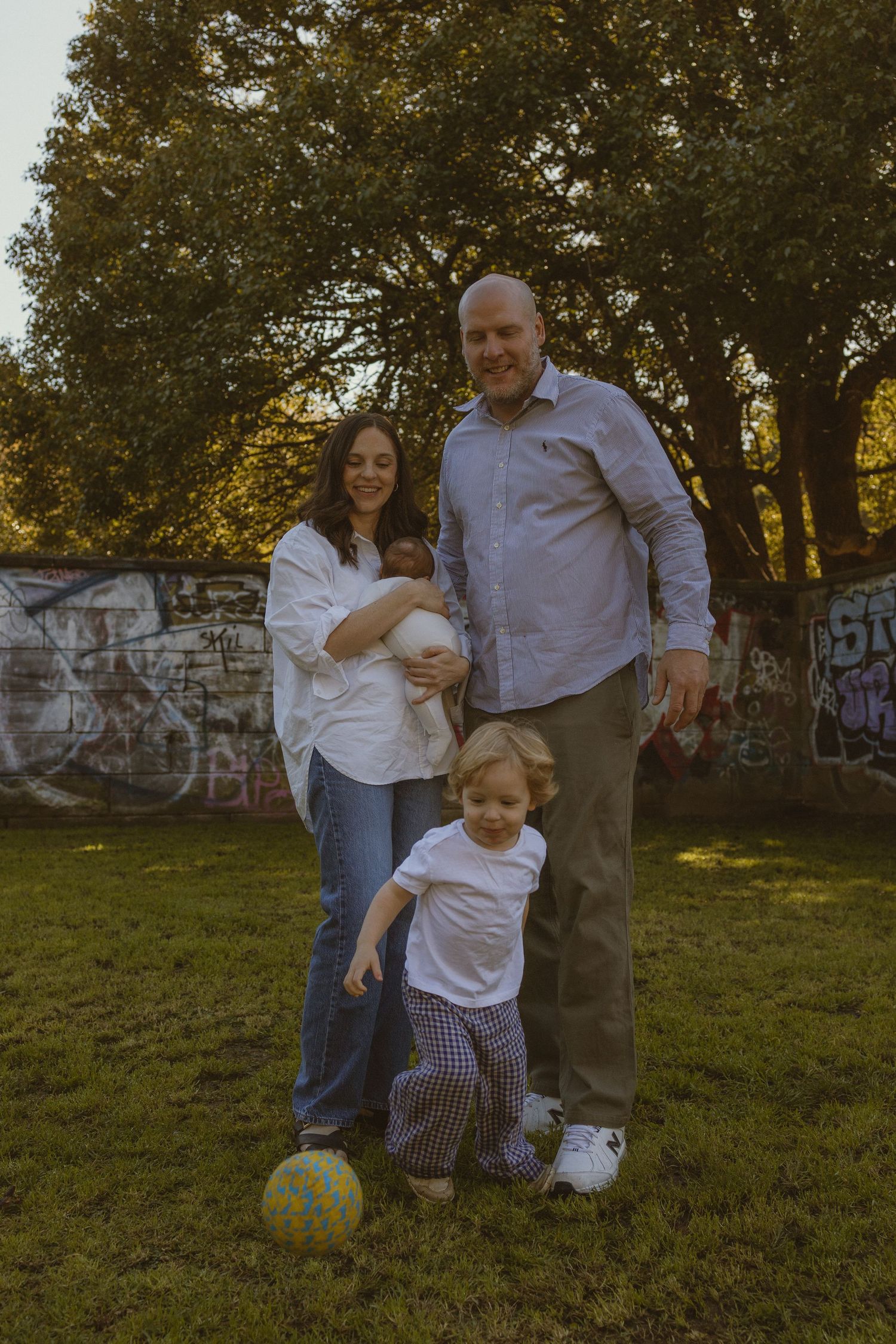 Family of four posing outdoors on grass, parents with newborn and toddler playing with soccer ball near graffiti wall.
