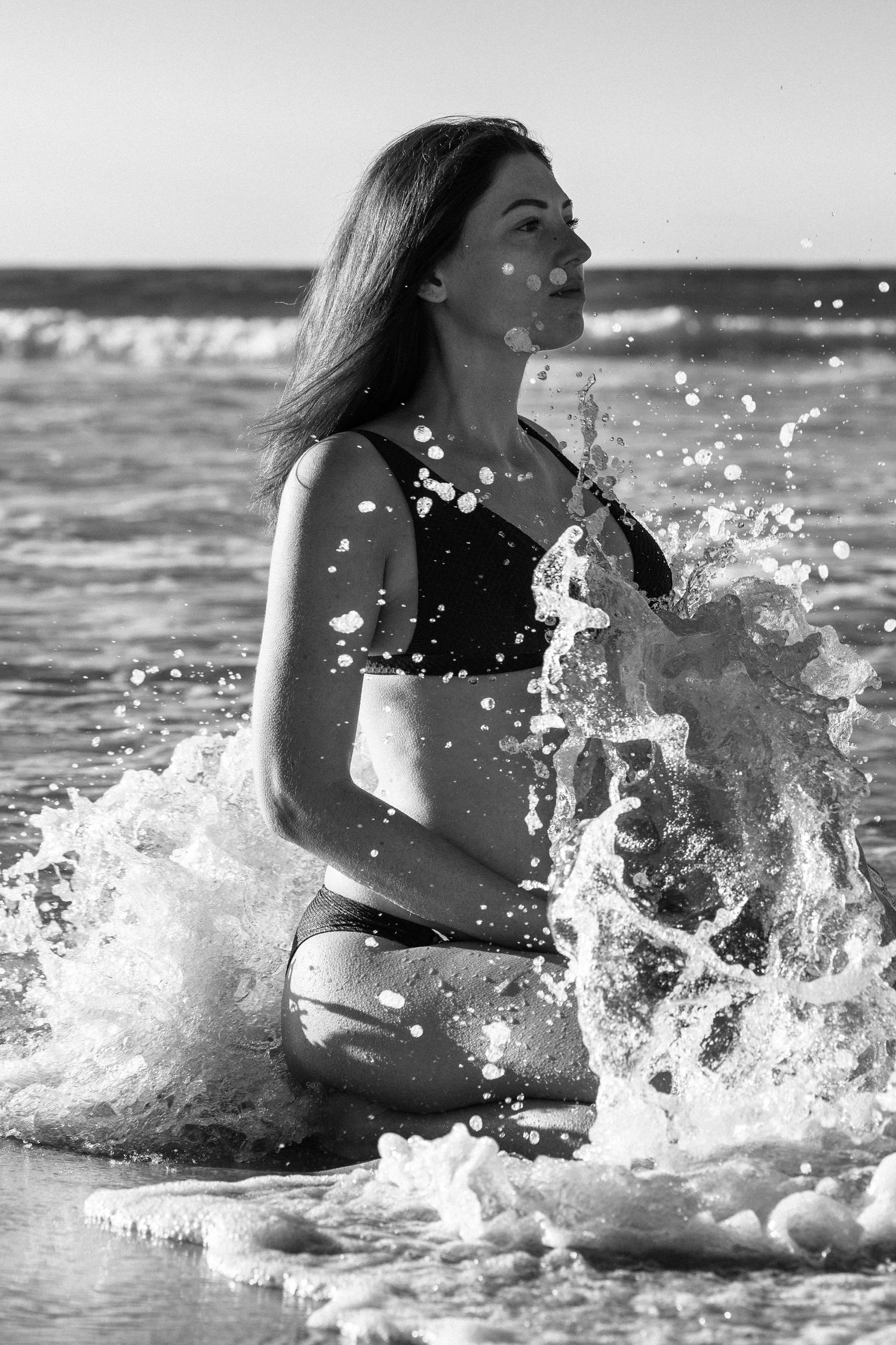 Black and white photo of a woman in a bikini sitting in ocean waves with water splashing around her on a sunny beach.