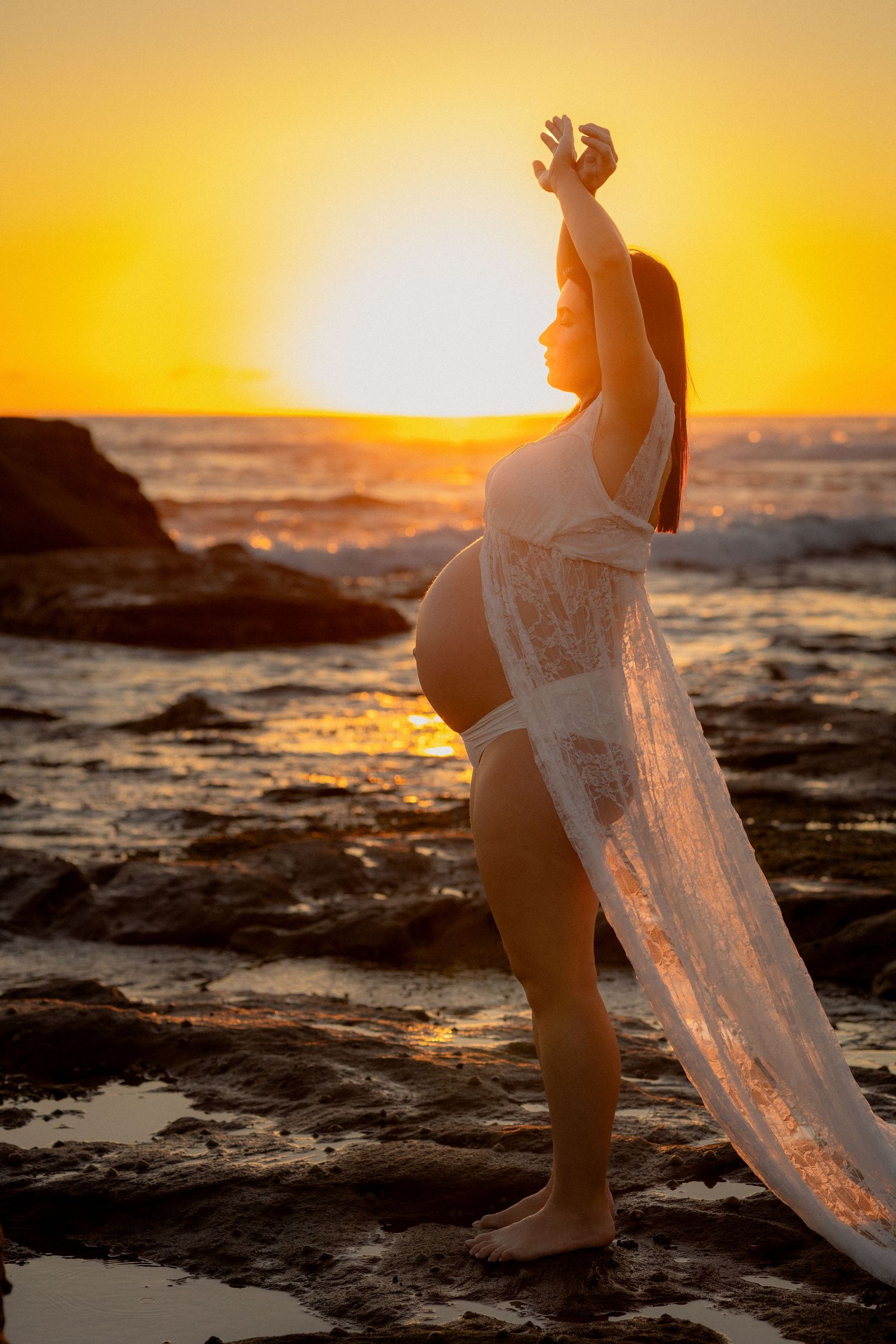 Pregnant woman in white lace wrap poses at rocky beach during golden sunset, silhouetted against vibrant orange sky.